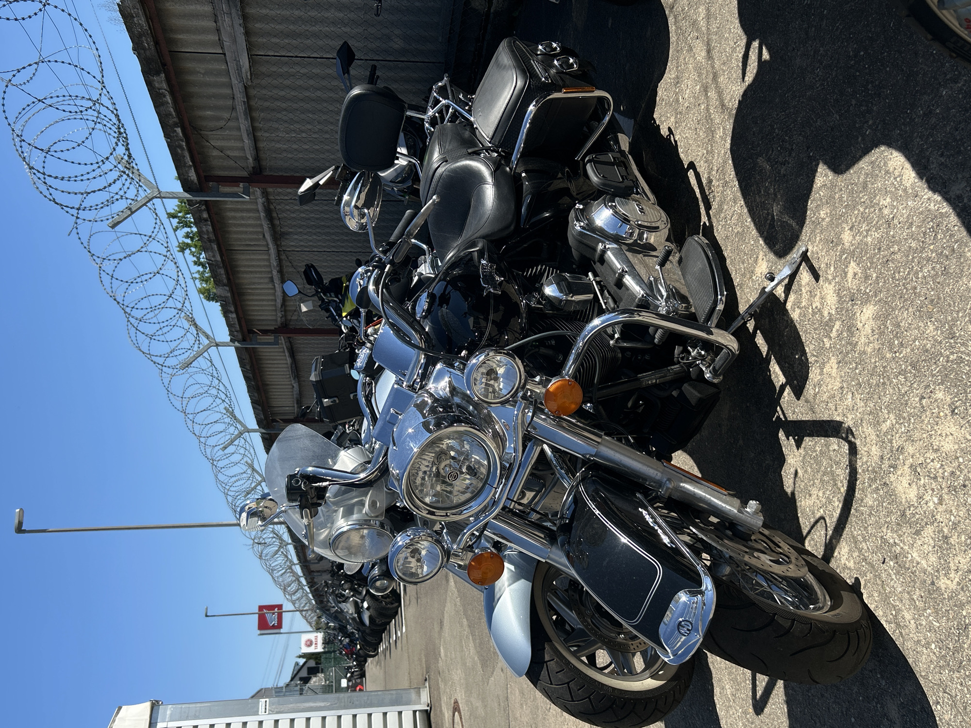 The image shows a row of motorcycles parked in what appears to be an outdoor storage or sales area. The motorcycles are predominantly black and chrome, with classic cruiser styling. Barbed wire is visible along the top of a building or structure in the background. Flags of Yamaha and Honda can be seen. Based on the presence of these flags, it is likely in the area of Bernhausen, Germany.