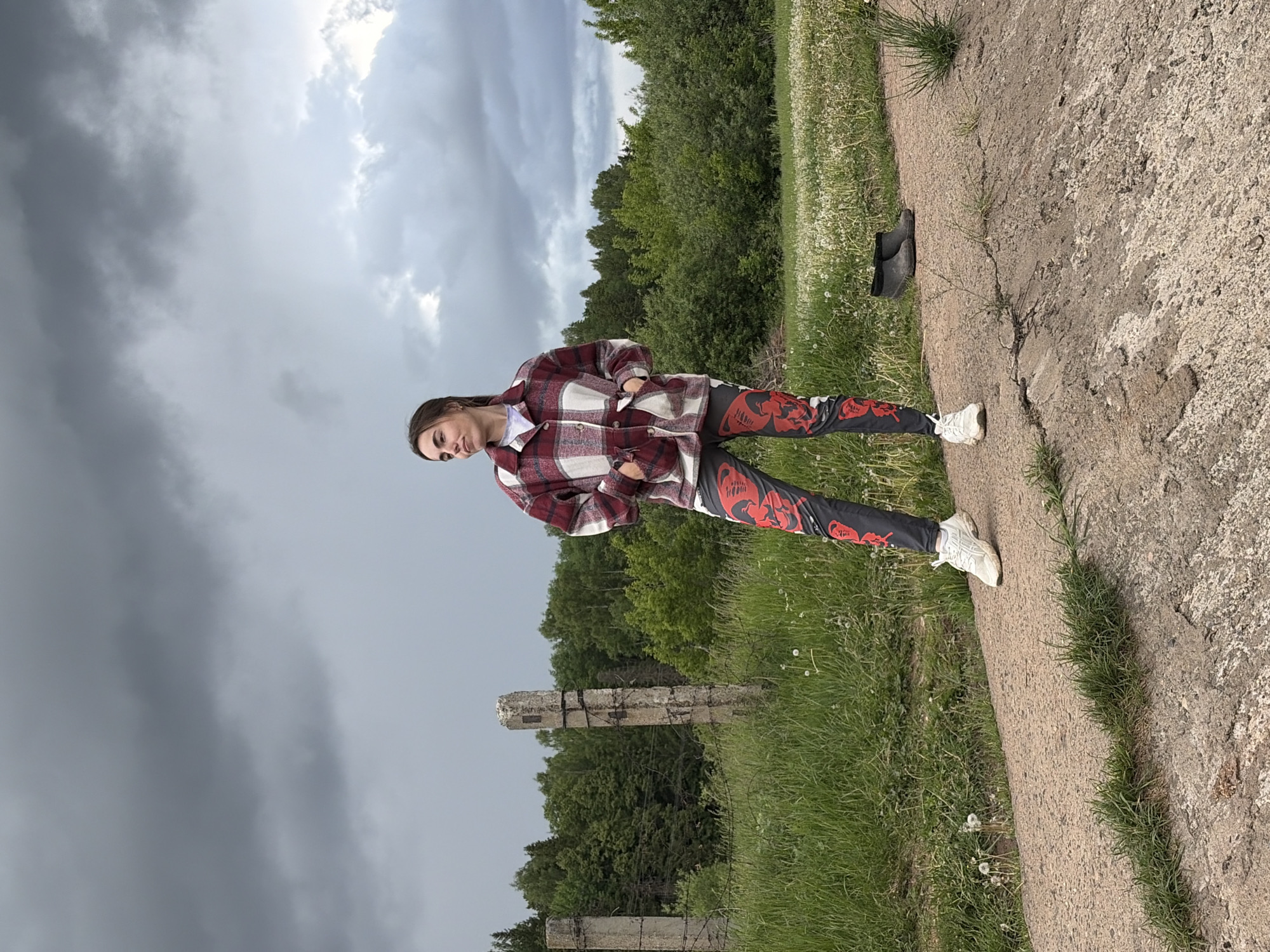 A young woman stands outdoors with a grey, overcast sky above. She wears a maroon and white plaid jacket over a white shirt, black leggings with a red design, and white sneakers. The environment includes trees and grass with dandelions. A cracked concrete edge is visible, bordering the grass. Two black shoes are discarded further along the concrete edge. In the distance are the remnants of broken concrete structures entwined with barbed wire. The location is Nizhnii Vishur, Russia.