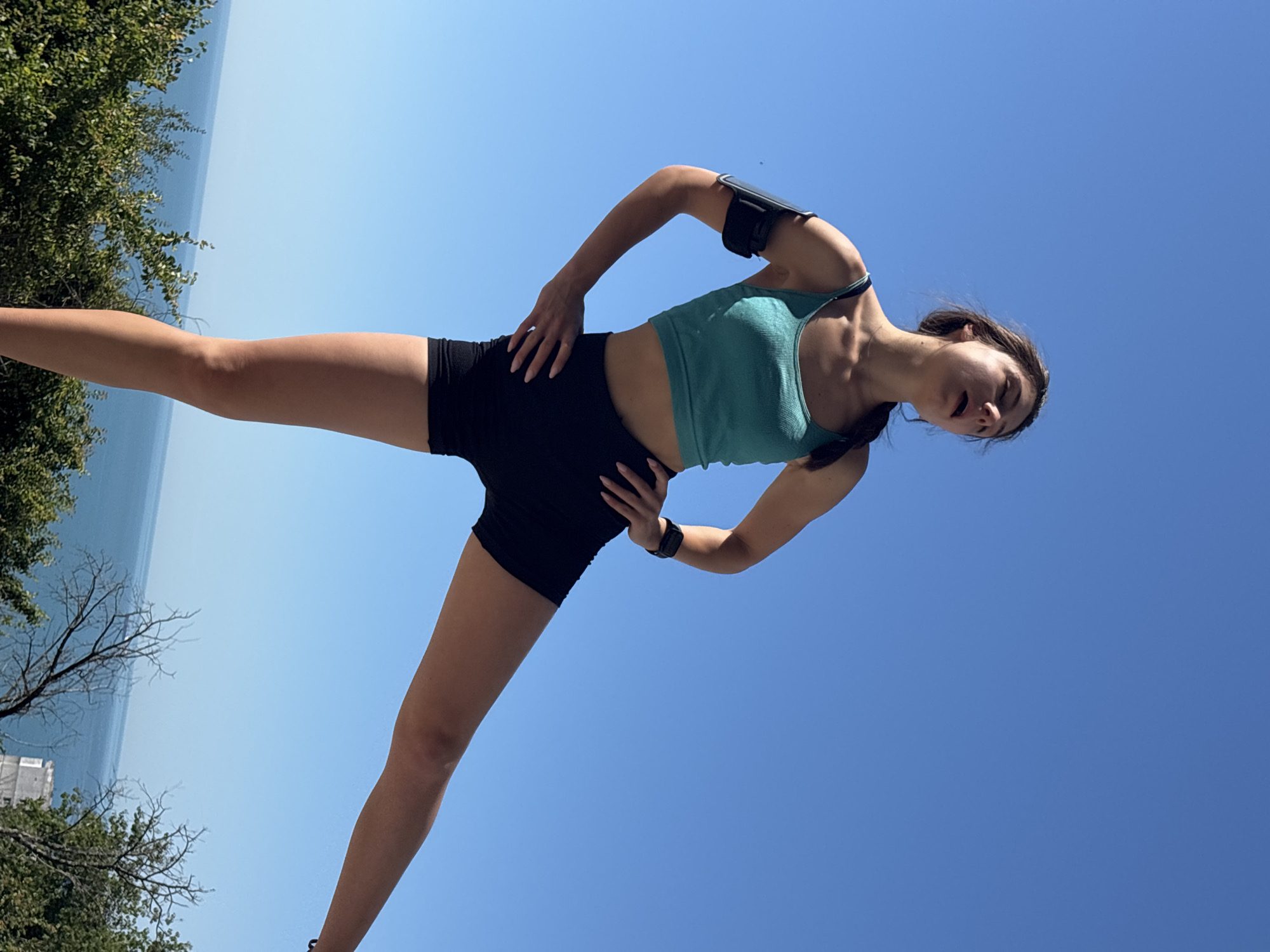 A young woman with brown hair braided down her back is performing an exercise outdoors. She is wearing a teal tank top and black shorts, with an armband on her left bicep. Her right leg is extended to the side, and her head is tilted back with her eyes closed, suggesting exertion or deep breathing. She is positioned against a clear blue sky, with a glimpse of the sea and green foliage below. The scene is likely set in a scenic, elevated location within Varna, Bulgaria, perhaps along its coastline or a park offering panoramic views.