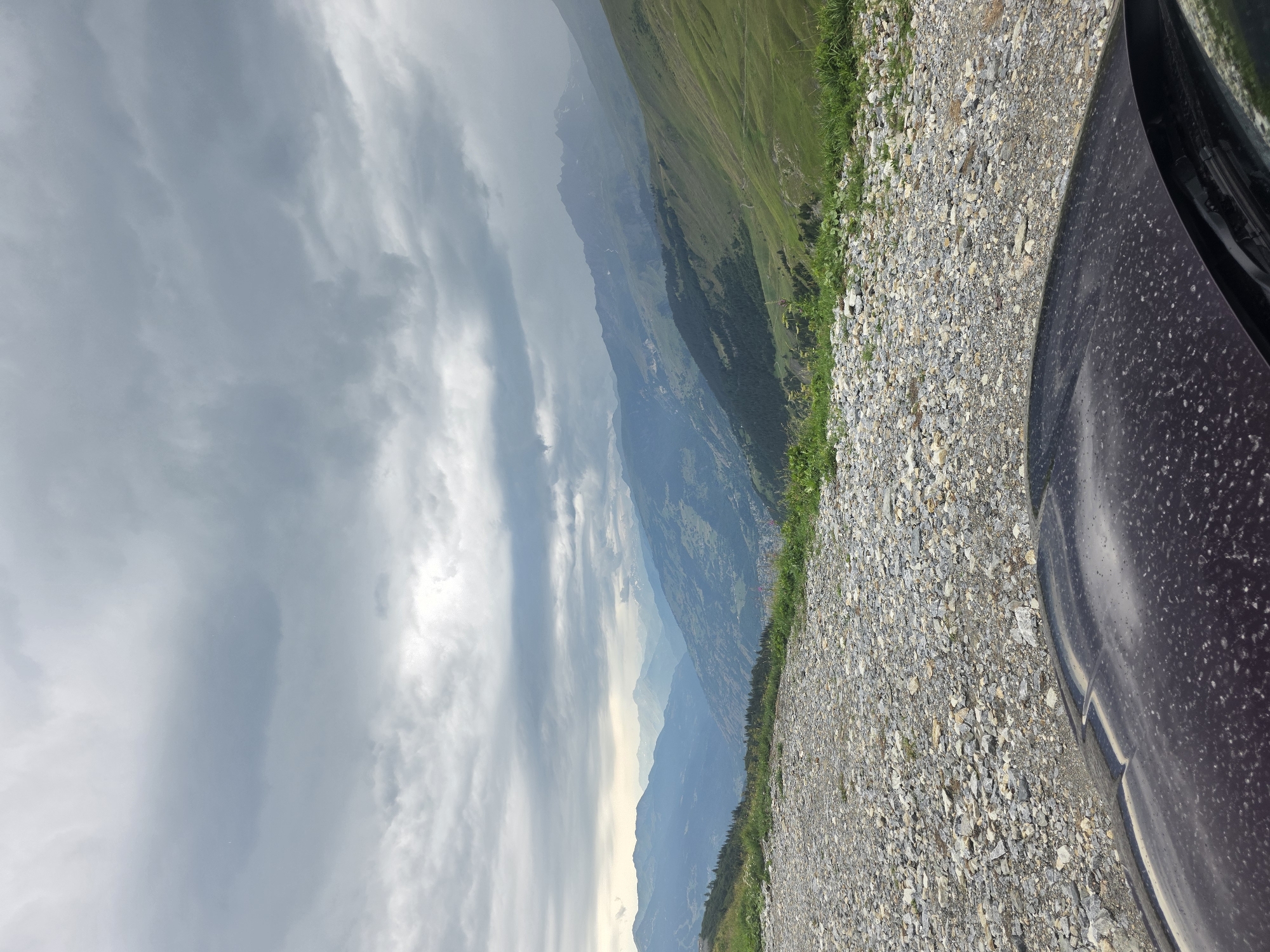 An overcast sky looms over a mountainous landscape. In the foreground, the hood of a dark-colored vehicle is visible, speckled with dust. Beyond the vehicle, a gravel road curves into the distance, bordered by green grass and wildflowers. The road leads to a panoramic view of a valley with a town nestled within, surrounded by rolling hills and majestic mountains that recede into the hazy distance. The scene appears to be a scenic overlook in the vicinity of Séez, France. There are no people visible in the image.