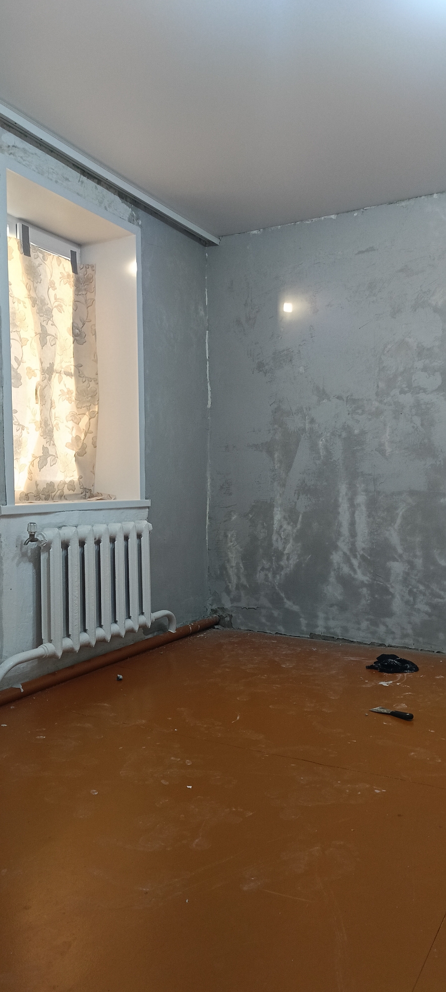 The image depicts an interior view of a room, likely undergoing renovation or preparation for finishing work. The walls are a rough, gray plaster finish, with visible imperfections and variations in texture. A section of the wall in the foreground, near the corner, shows a distinct bright square light source, possibly a recessed ceiling light or a reflection. To the left, there is a window with a light-colored curtain patterned with floral motifs. Beneath the window sits a white radiator, with visible pipes extending to the left. The floor is a polished, reddish-brown surface, appearing somewhat dusty or smudged with white marks. In the lower right portion of the image, a black cloth or garment is visible on the floor, alongside a putty knife or scraper with a black handle. The ceiling is white and appears to be a suspended ceiling system, with a visible metal track along the perimeter near the walls. The overall environment suggests a domestic or residential space in a state of disarray or transition. The location is identified as Хохлы, Russia, but this information cannot be verified from the visual content alone.