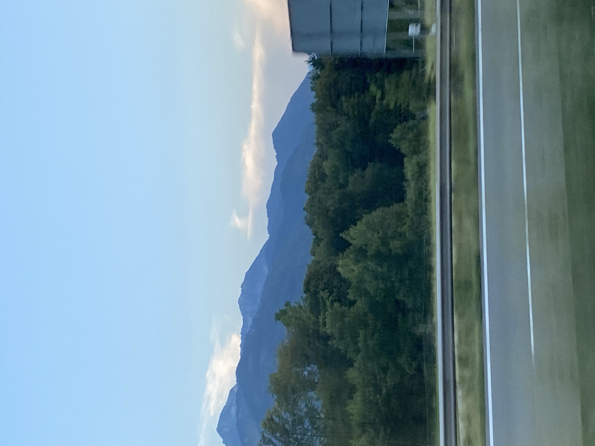 The image shows a scenic view from a highway in Grabenstätt, Germany. In the foreground, a section of the highway with white lane markings is visible, along with a guardrail and a grassy shoulder. Beyond the highway, a dense forest of green trees stretches across the midground. In the background, majestic blue mountains rise against a pale blue sky with wispy clouds. A large gray sign structure is partially visible on the right side of the image. The scene appears to be captured during daylight, likely in the late afternoon or early evening.
