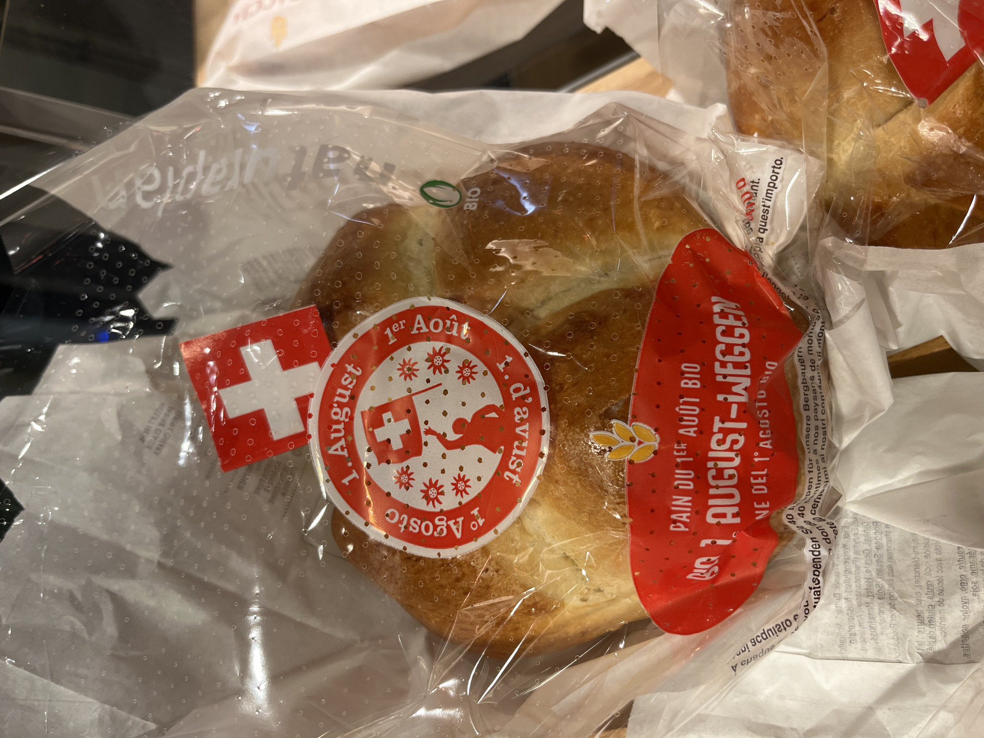The image shows a close-up of a packaged sweet bread product, commonly known as "1er Août Weggen" or "1er Août Brot," a traditional Swiss pastry made for Switzerland's National Day on August 1st. The bread is golden brown and braided, enclosed in a clear plastic bag. Two decorative stickers are affixed to the bread. The top sticker is round with a red and white Swiss flag and the text "1. August" and "1er Août" in a festive design. The bottom sticker is also red and white, featuring a wheat stalk and the text "PAIN DU 1er AOÛT BIO" and "1 AUGUST-WEGGEN" in bold lettering, along with "VE NE DEL 1° AGOSTO BIO" in smaller text. A small green "BIO" logo is visible on the plastic bag to the right of the top sticker. The bread is placed on a surface that appears to be a wooden table or counter. The background is blurred, showing some white packaging material and a hint of a dark surface, possibly shelves or another product. There are no people or identifiable landmarks visible in the image. The context suggests this product is being sold or displayed in a retail environment, such as a bakery or a grocery store. No specific location in the city of Geneva, Switzerland, can be identified from the visual information provided.