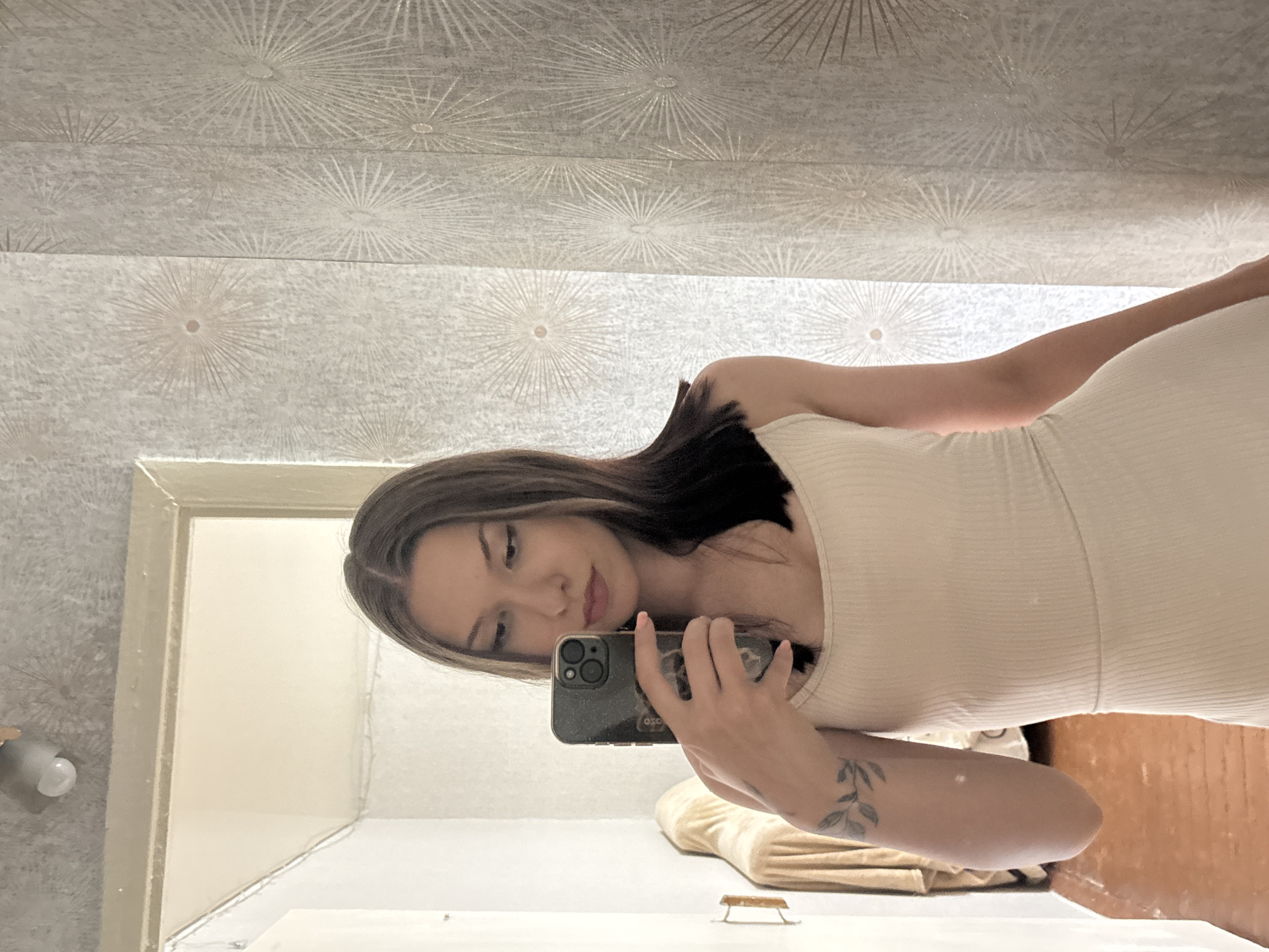 A young woman with long, dark brown hair and fair skin takes a selfie in a room. She is wearing a cream-colored, sleeveless dress. She is holding a dark-colored smartphone in her right hand, angled to capture her reflection. A tattoo of a leafy branch is visible on her left forearm. The room has light-colored walls with a textured wallpaper featuring a starburst pattern. To the left of the woman, a doorway leads to another area, where a piece of furniture covered in a light-colored cloth is visible. The floor is wooden. The image appears to have been taken indoors, possibly in a residential setting. There is no specific information to determine the location in the city of Inskoy, Russia.