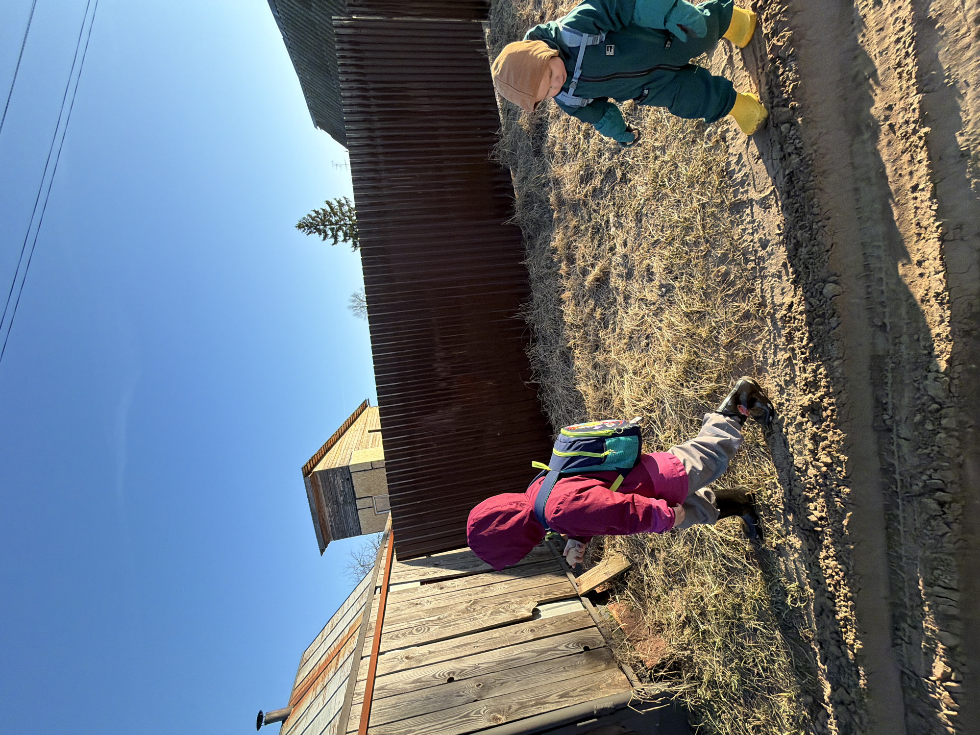 The image shows two children in outdoor clothing standing on a dirt path next to a building with corrugated metal siding in Chaikovsky, Russia. One child wears a red hooded jacket and a backpack, while the other wears a green jumpsuit and yellow boots. The sky is blue.