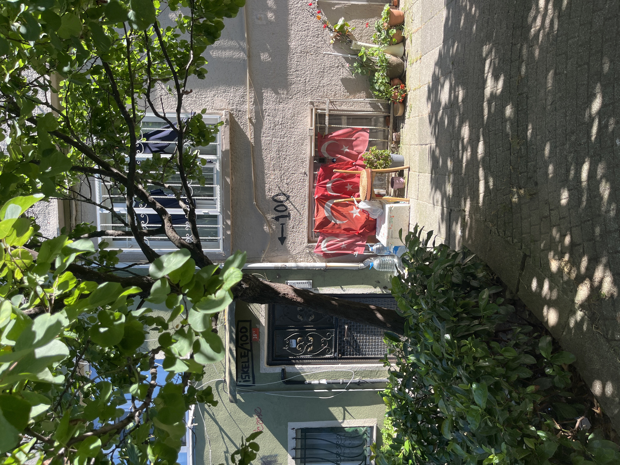 In Kadıköy, Turkey, on a sunlit day, a narrow sidewalk is visible in the foreground, paved with grey cobblestones and dappled with tree shadows. To the left, a dense green hedge lines the sidewalk, partially obscuring the lower part of a building.
The building, situated behind the hedge and a tree, has a light green facade on the left and a textured, cream-colored facade on the right. The left section features a window with a metal grate and a dark metal door adorned with decorative patterns. Above the door, a sign reads "İSKELE/100" with a smaller red number "16" below it. To the right of the door, a black arrow points towards the number "100" painted on the cream-colored wall.
The cream-colored section of the building has a large window with a metal grate, behind which dark fabric is visible. In front of this window, several Turkish flags with red backgrounds and white stars and crescents are displayed, hanging from a horizontal bar. A simple wooden chair with a woven seat is placed in front of the flags, and next to it are two plastic water bottles and a white rectangular object.
Further to the right, a collection of potted plants with red and green foliage, as well as a white vase with flowers, are arranged along the base of the building. The scene suggests a residential area, with decorative elements and everyday objects arranged outside.