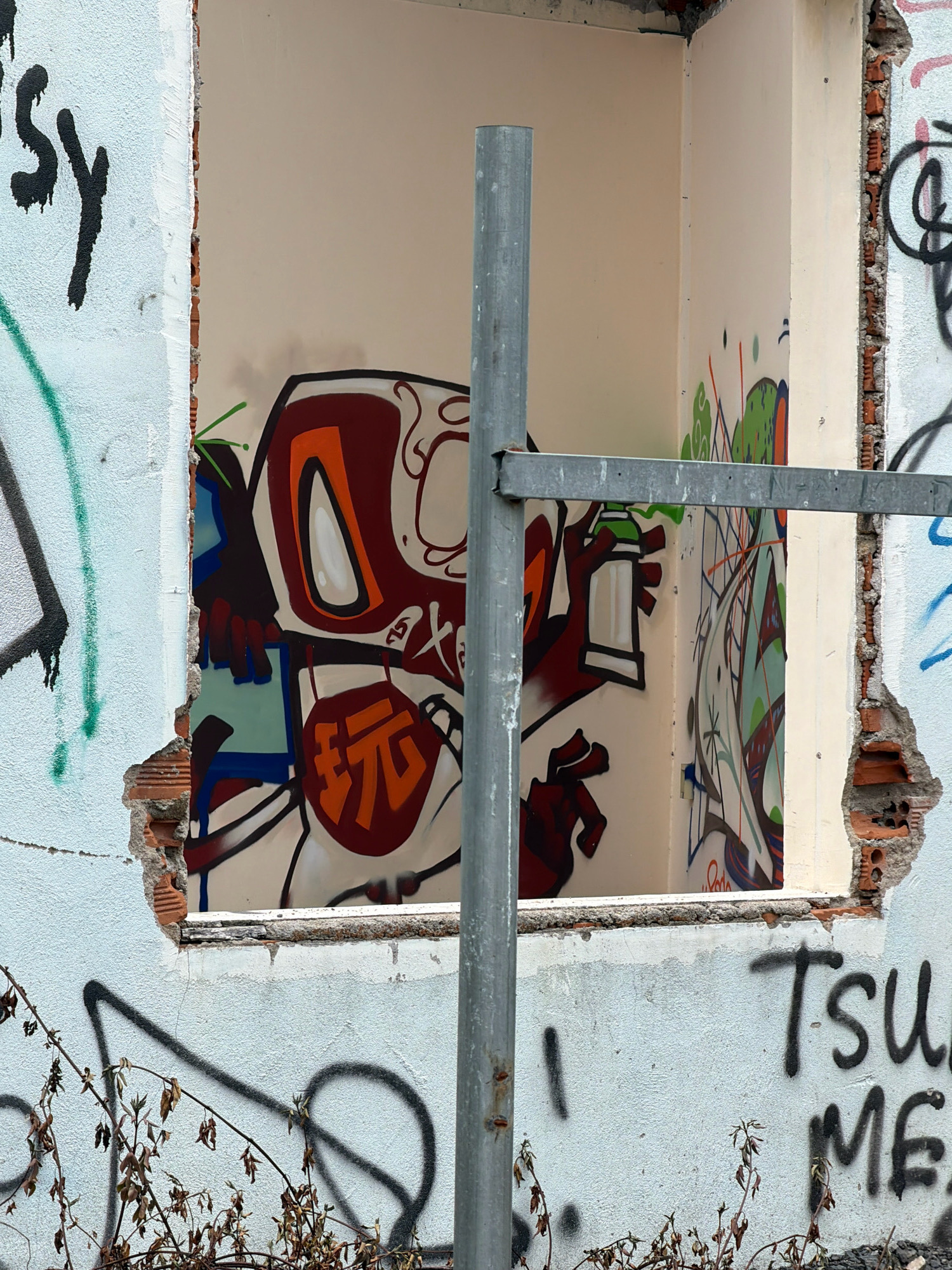 The image captures a section of a dilapidated white building with exposed brickwork and graffiti. A large, rectangular opening in the wall reveals an interior space decorated with colorful graffiti art. The art features cartoon-like characters with bold outlines and vibrant colors, including a prominent figure with a red and orange head and large eyes. Various abstract shapes and lines in green, blue, and red are also visible.
A vertical metal pole stands in front of the opening, partially obscuring the graffiti. A horizontal metal bar is attached to this pole and extends to the right, suggesting scaffolding or a temporary support structure.
The exterior wall of the building is also covered in graffiti. To the left, black lettering, possibly "SY," is visible with dripping paint. To the right, black spray-painted text reads "Tsu" and "ME."
The ground in the foreground is partially covered by dry, dead vegetation and debris. The overall scene suggests an abandoned or under-construction site within a city.
Based on the provided information, this location is in Nha Trang, Vietnam.