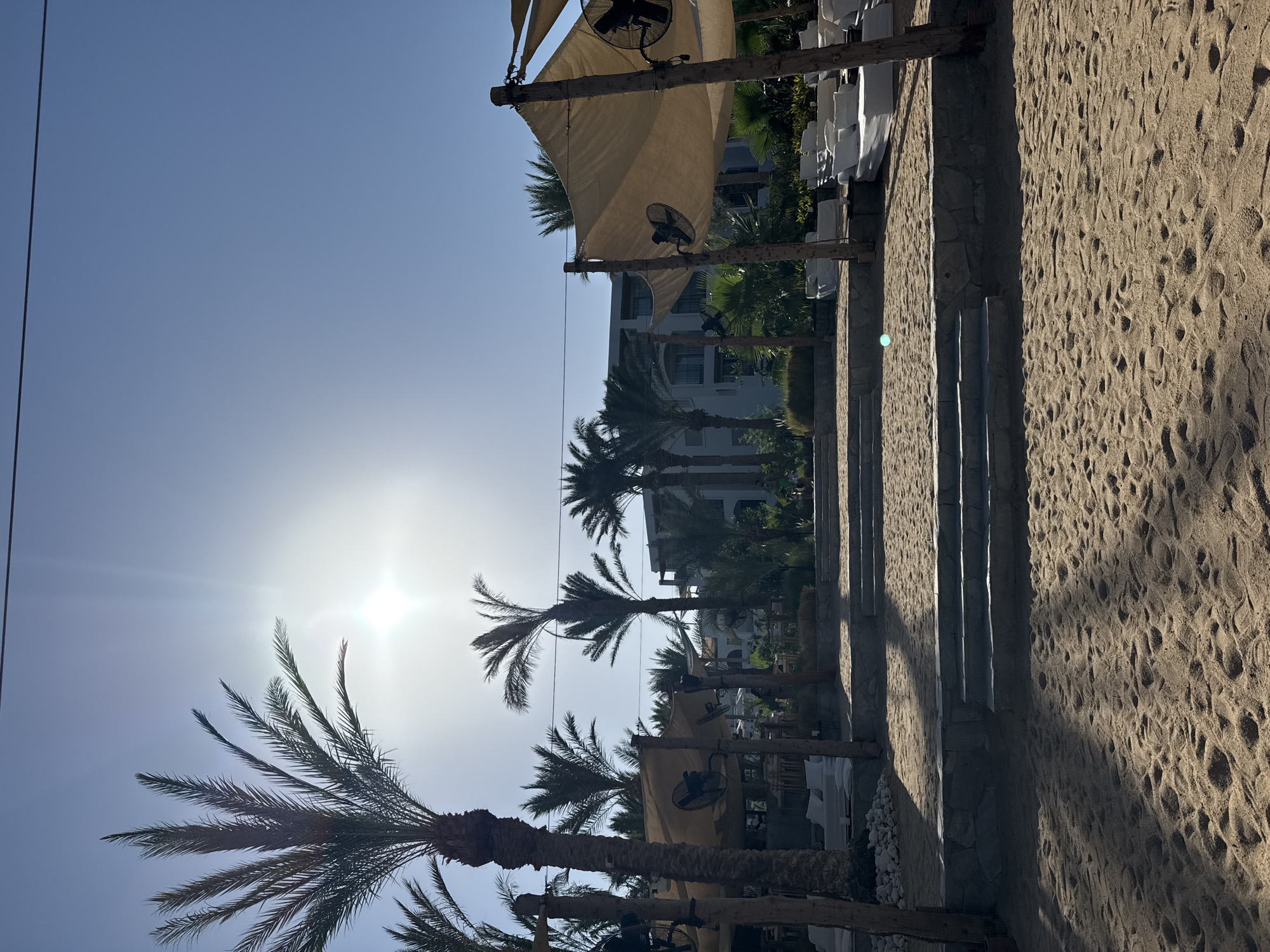 This image depicts a bright, sunny day at a beach resort in Hurghada, Egypt. The scene is dominated by a clear blue sky with the sun shining intensely, creating lens flare. In the foreground, a sandy beach stretches along the right side of the frame, marked by footprints and pebbles. A stone wall with vertical pillars runs parallel to the beach, casting long shadows across the sand.
To the left, several palm trees are visible, some silhouetted against the bright sky. Behind the palm trees, there is a white building, likely part of the resort, with balconies and windows. Overhanging canvas awnings, supported by wooden beams, are stretched over seating areas, and electric fans are attached to the beams, suggesting a focus on comfort in the warm climate. White towels or fabrics are hanging neatly on a rack to the left of the stone wall.
The image captures a tranquil atmosphere, with no people visible. The time of day is likely late morning or early afternoon, given the harsh sunlight and long shadows. The overall impression is one of a well-maintained resort area designed for relaxation and enjoyment under the Egyptian sun. No visible text is present in the image.
