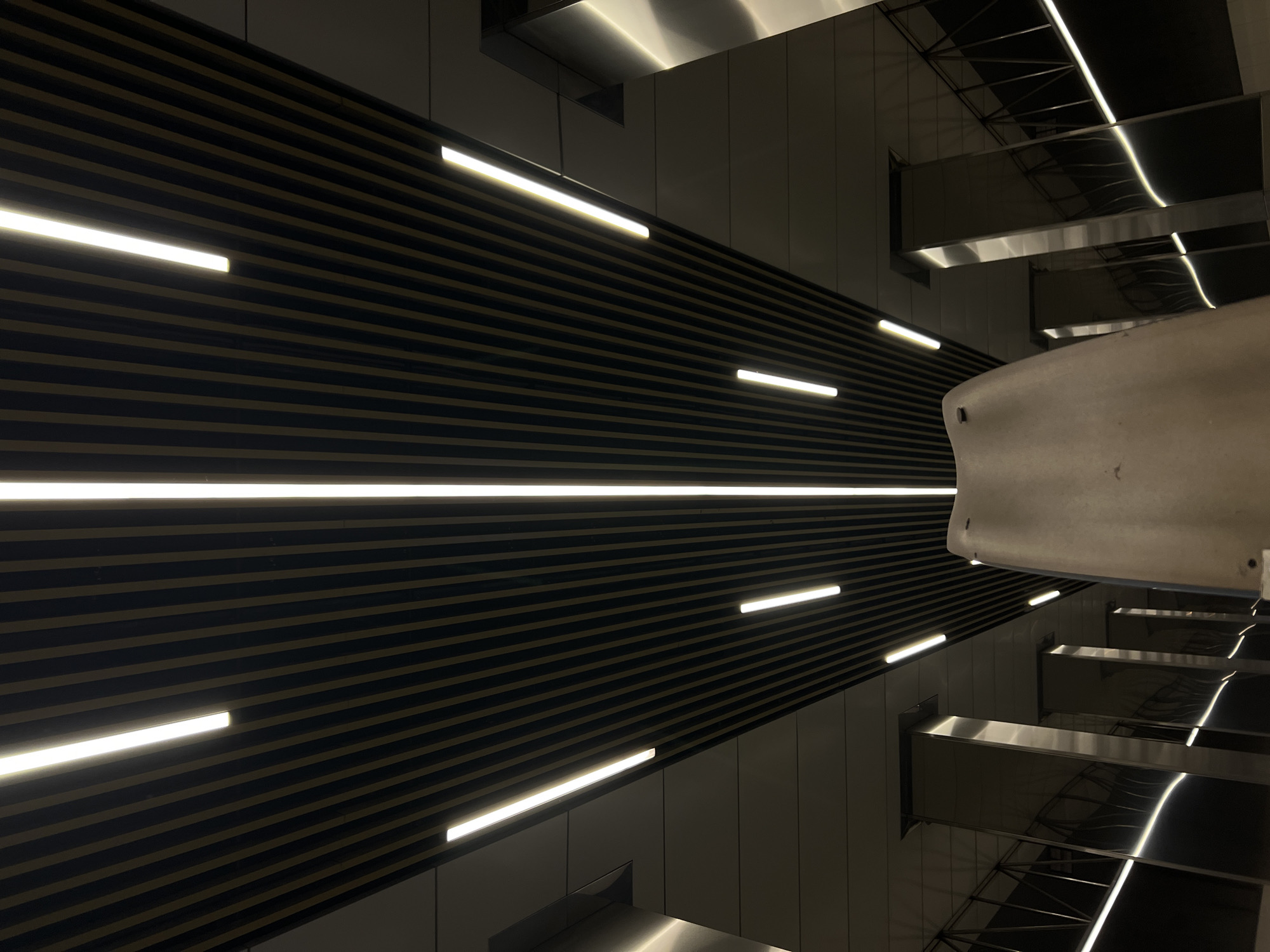 The image shows an interior architectural scene, likely a modern transportation hub or public space, in Moscow, Russia. The perspective is tilted upwards, emphasizing the ceiling's design.
The ceiling features dark, horizontal striped panels interspersed with linear LED light fixtures, creating a dynamic pattern of light and shadow. To the right of the frame, there are metallic or reflective architectural elements, possibly structural beams or components of a staircase. The lighting is diffused and creates specular highlights on the metallic surfaces. The overall impression is of a sleek, contemporary design.