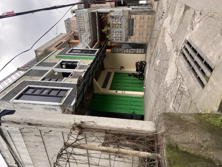 The image shows a narrow street in Margaasih, Indonesia (latitude: -6.961, longitude: 107.546), lined with two-story concrete buildings. The street slopes downhill, with a drainage grate visible in the foreground. A motorcycle is parked in front of a green garage door, and a tall concrete wall with a metal fence is visible on the left side of the image. An overhead power line runs along the buildings. There is no one visible in the image.