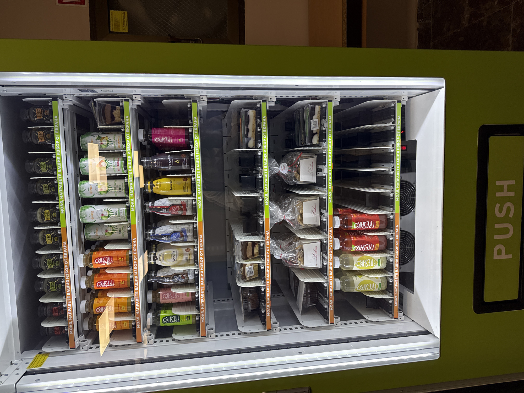 A vending machine is shown in a medium shot. The machine is stocked with an assortment of drinks and packaged goods. The left-most column contains multiple bottles of "FRESHKO" drinks, followed by bottles of fruit-flavored coconut water, and then a selection of vanilla, mocha, and dark coffee. The next column contains wrapped pastries, cakes, and cookies. Finally, the right-most column contains more varieties of "FRESHKO" drinks. The word "PUSH" is printed on the right side of the machine in bold green letters. The sides of each column say, "МОЛЯ, НАПРАВЕТЕ ВАШИЯ ИЗБОР ОТ ЕКРАНА". The location of the image is Unknown, Unknown.