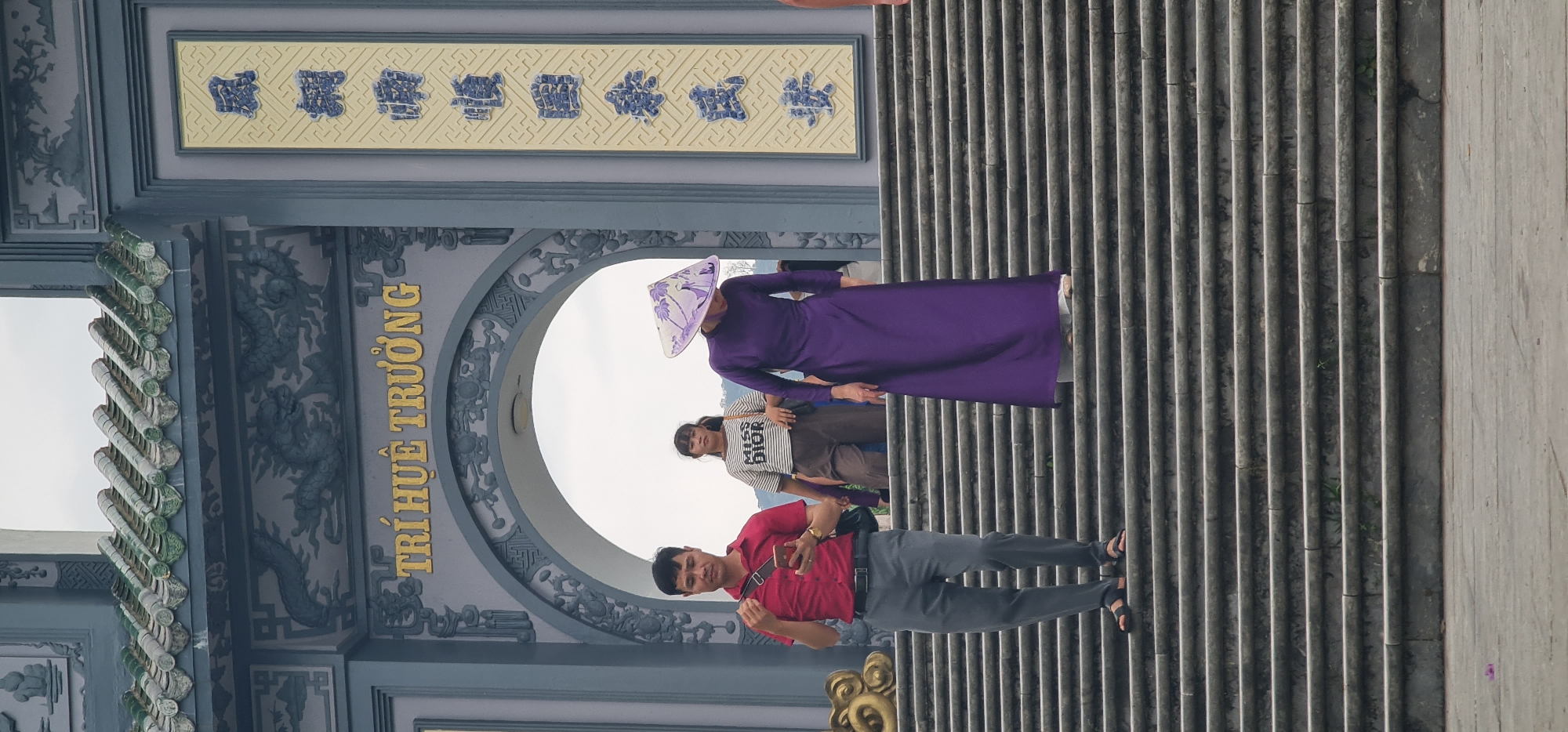 The image displays a vibrant scene at an ornate architectural site, likely a temple gate or archway, in Đà Nẵng, Vietnam. The sky appears overcast and bright, suggesting daytime.
Three people are visible on a set of wide, ribbed stone steps leading up to the archway. In the foreground, a man wearing a red polo shirt, grey trousers, and black sandals stands smiling and looking towards the camera, holding a smartphone. Behind him, to his right, is a young woman dressed in a striped t-shirt with "MISS DIOR" printed on it and brown pants, looking slightly to the side. Next to her, a woman in a traditional long purple áo dài and a patterned conical hat (nón lá) stands facing forward.
The prominent feature in the background is a large, elaborate grey archway. Its columns and arch are adorned with intricate relief carvings of what appear to be dragons and floral motifs. Along the vertical beam of the archway, golden Vietnamese text is clearly visible, spelling out "TRÍ HUỆ TRƯỜNG". Above the arch, a horizontal decorative panel features six blue characters, likely Chinese or Hán Nôm, set against a yellow, patterned background. The roof structure of the archway is embellished with decorative green-tiled elements. The overall impression is a cultural or historical landmark, with visitors admiring or posing at the site.