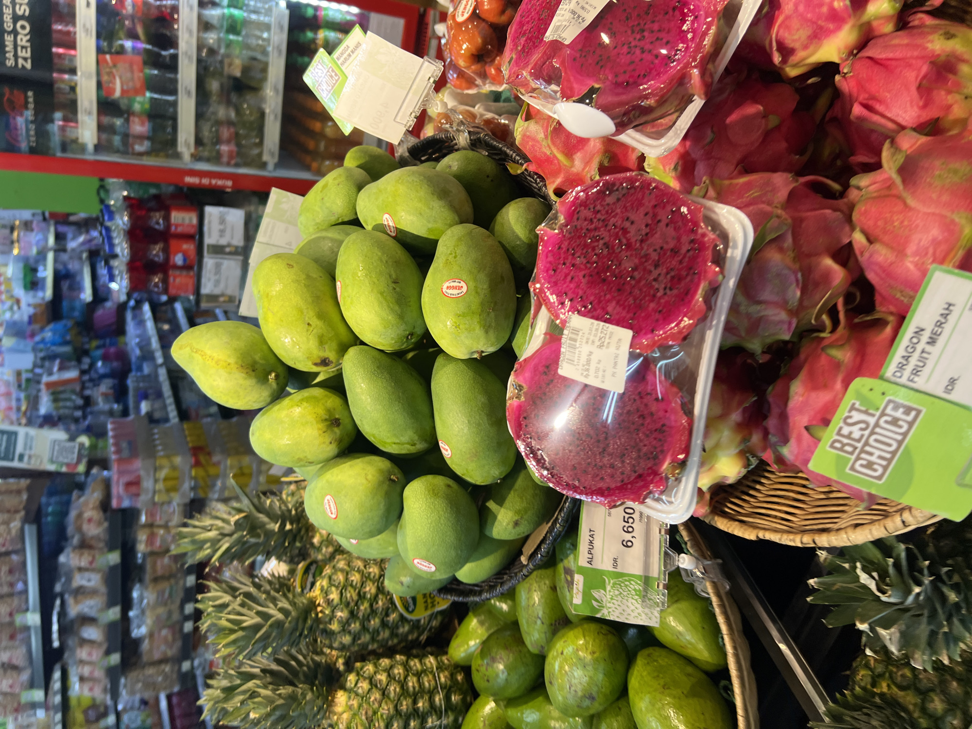 The image depicts a brightly lit produce section of a supermarket in Kuta, Indonesia. Several varieties of fruits are displayed in baskets and on shelves. In the foreground, a basket overflows with green mangoes, some of which have red stickers on them. To the left of the mangoes, a collection of green avocados is visible. Adjacent to the mangoes, two halves of a sliced pink dragon fruit are presented in clear plastic packaging. Behind the mangoes, several pineapples are stacked. To the right of the dragon fruit, a pile of whole pink dragon fruits is arranged. In the background, shelves are stocked with various packaged goods and beverages, including drinks in red bottles and bottles in a clear refrigerator. A price tag for avocados reading "IDR. 6,650" is visible in the lower left portion of the image. Another price tag for dragon fruit is partially visible in the lower right. A green sign that says "BEST CHOICE" is placed in front of the dragon fruit.