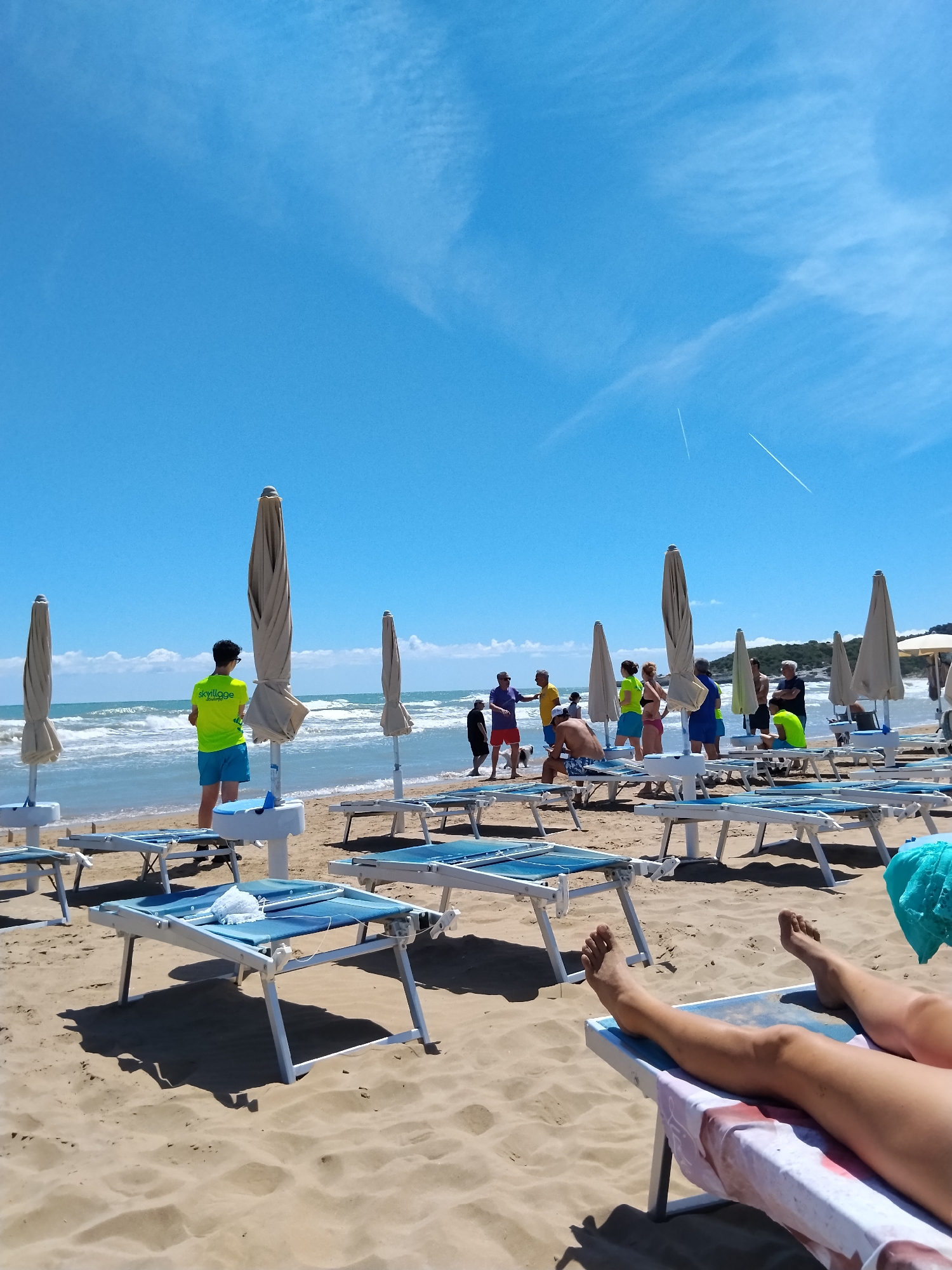This image captures a sunny day at the beach in Peschici, Italy, likely during the summer season. The scene is dominated by a clear blue sky with wispy clouds and two faint contrails from airplanes. The foreground is filled with rows of unoccupied, blue-and-white beach loungers with some towels neatly placed. In the mid-ground, a group of people are gathered near the water's edge. Among them, a person wearing a bright yellow shirt with the logo "Skyvillage" and blue shorts stands facing the ocean. Further back, several closed beach umbrellas are lined up on the sand. The ocean in the background exhibits moderate waves. The sandy beach is clean and well-maintained, with shadows cast by the loungers and umbrellas, indicating a strong sun. The overall atmosphere is one of leisure and vacation.