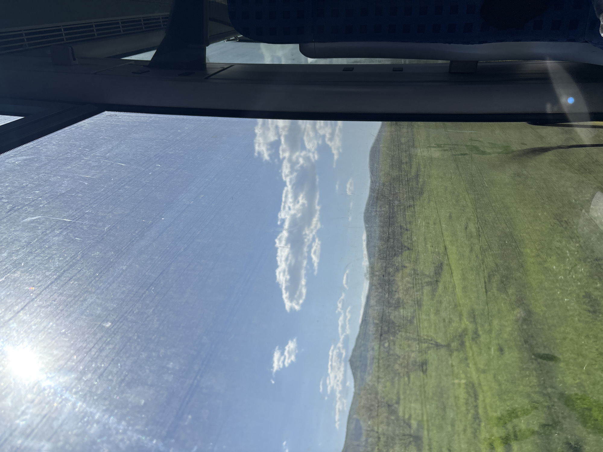 The image appears to be taken from inside a moving train or bus, looking out the window. The foreground is dominated by the dirty glass window reflecting the sky and clouds on the left, and a field on the right. The sun's reflection is visible on the lower left. Above the window, parts of the vehicle's interior are visible, including a blue headrest. The field appears to be an agricultural field, with clear rows of crops. It is difficult to determine the exact location within Ovchepoltsi, Bulgaria without further information. There is no explicit evidence of people, subjects, objects, actions, or interactions other than the view of the passing landscape.