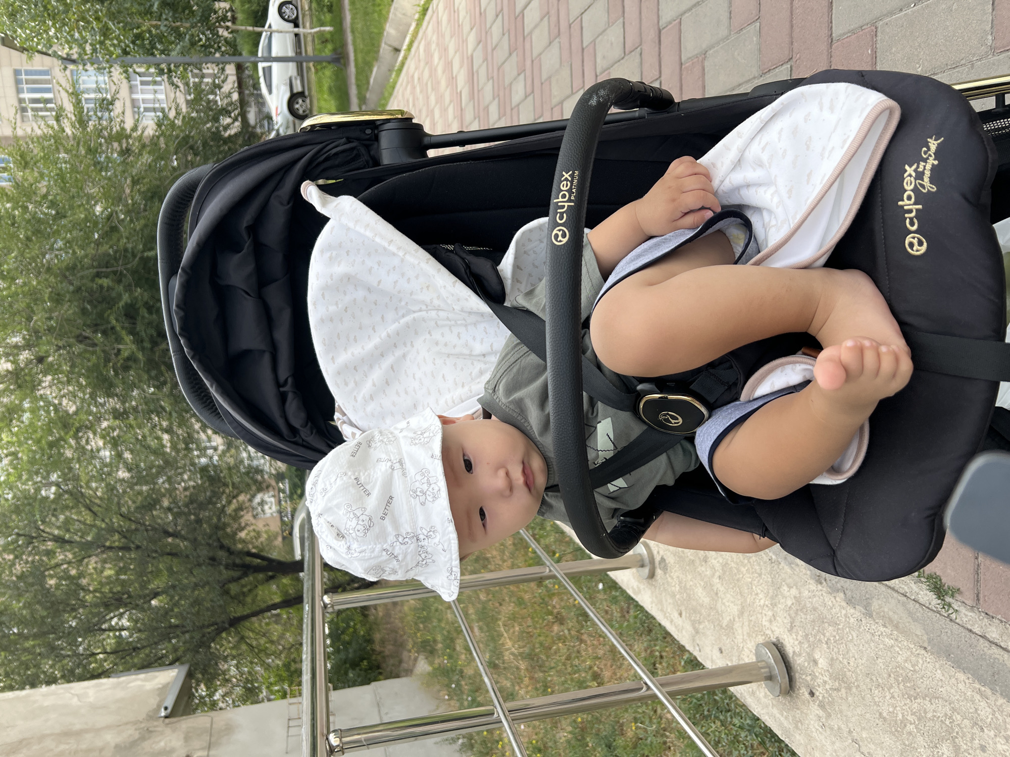 A young East Asian child, wearing a white bucket hat with a pattern of teddy bears and the word "BETTER" repeated, sits in a black Cybex Platinum stroller. The child is dressed in a green t-shirt and gray shorts. The stroller has a white canopy with a cloud pattern and a gold trim. The child's legs are visible, with one foot resting on the stroller's seat. The background shows a building with windows, trees, and a sidewalk made of red bricks. A white car is parked on the street in the background. The scene appears to be in an urban environment, possibly in Bishkek City, Kyrgyzstan.