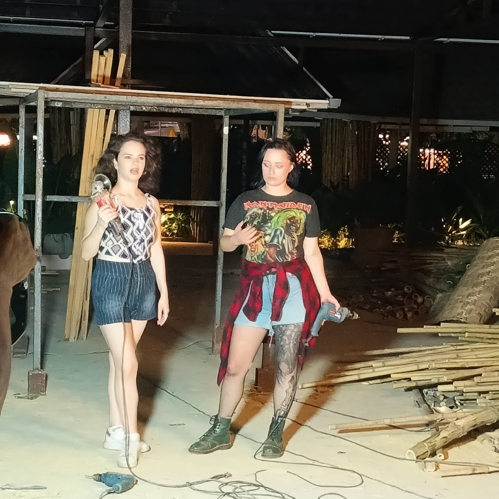 In Panaji, India, two women stand amidst construction materials, including bamboo and lumber, under the dimly lit frame of a partially constructed structure. The woman on the left wears a patterned top and striped shorts, holding a grinder, while the woman on the right is clad in an Iron Maiden t-shirt, denim shorts, and a flannel shirt tied around her waist, holding a drill; she has a tattoo on her left leg. Electrical cords from power tools are visible on the ground. The environment suggests active construction or renovation at night.