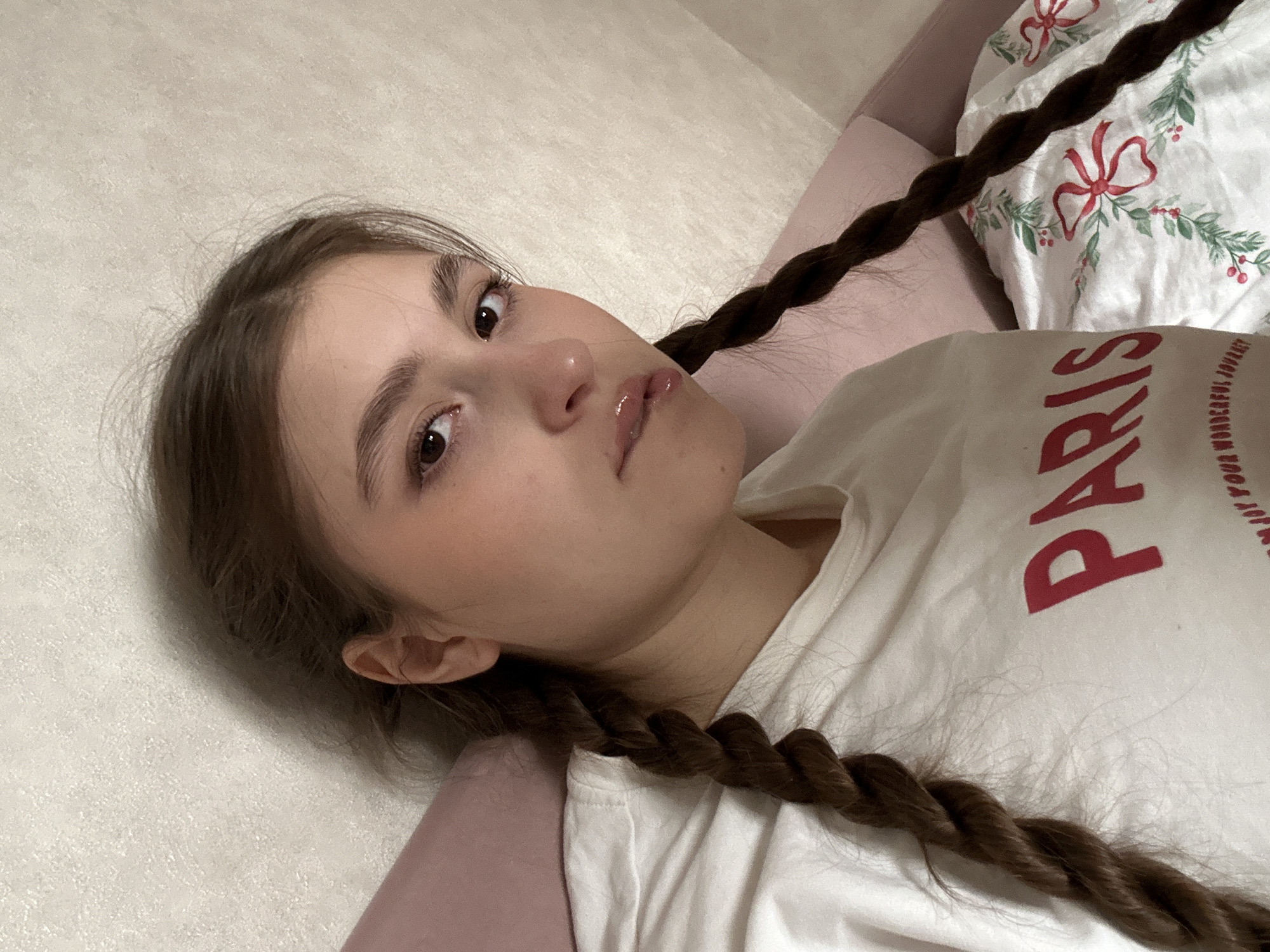 An indoor, high-angle portrait displays a female subject in what is likely a residential bedroom in Sarapul, Russia. The subject, with fair skin and dark brown hair, is styled with two braids resting on her chest and shoulders. She has dark eyes and glossy lips, with a direct gaze towards the viewer. She is wearing a white short-sleeved t-shirt, which has the word "PARIS" printed in red across the chest, with further red text partially visible below. The subject is positioned supine, with her head resting on a pink pillow. To her right, a white comforter or sheet featuring a pattern of green leaves and red bows is visible. The background consists of a light-colored, textured wall.