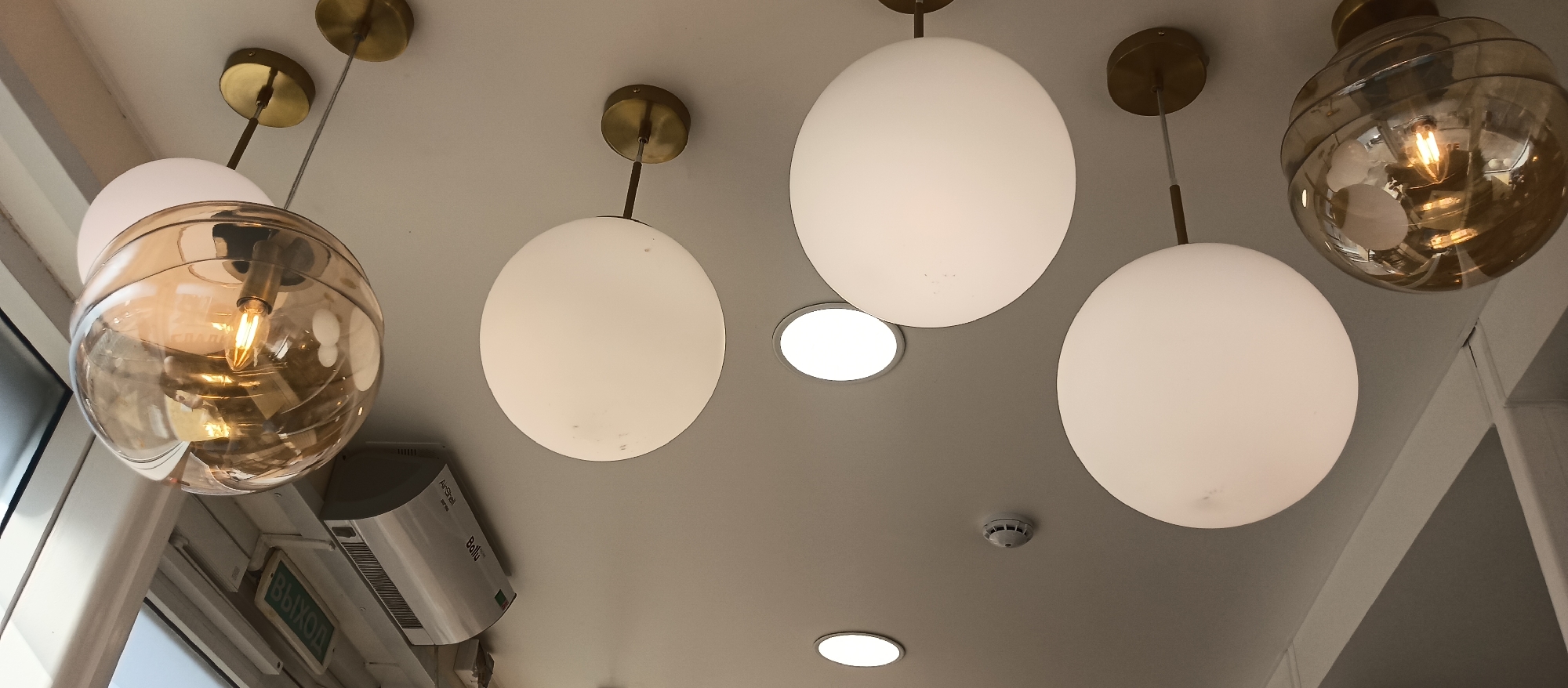 The image depicts a view of a ceiling with a series of modern lighting fixtures. There are four prominent globe-shaped pendant lights, with brass or gold-colored ceiling mounts. Two of these globes are partially translucent and have a golden hue, revealing their interior bulbs. The other two are opaque white, diffusing the light. The ceiling is white or light-colored. Two inset circular ceiling lights are also visible. A smoke detector is positioned centrally in the view. At the edge of the view, there's a corner with window and an exit sign, an air conditioning unit. The location appears to be indoors in a commercial or public setting.