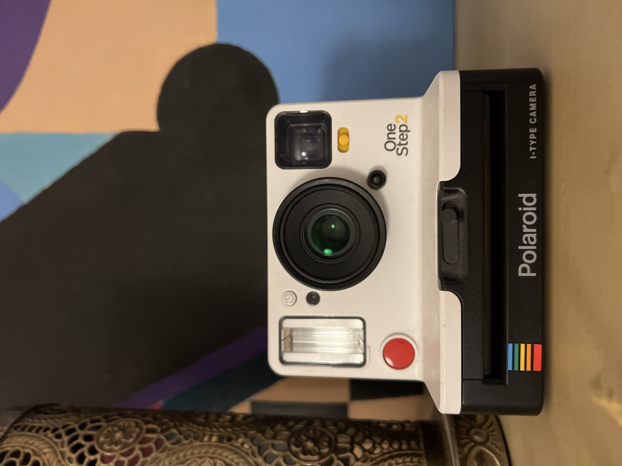 A white Polaroid OneStep 2 i-Type camera is positioned centrally in the foreground, facing the viewer. The camera is placed on a wooden surface, possibly a shelf or table, which occupies the bottom portion of the image. To the left of the camera, a decorative brass or gold-colored metallic object with intricate patterns is partially visible.
Behind the camera, the background consists of a wall with abstract, colorful geometric shapes. Large areas of black, peach, light blue, and purple are present in the artwork. The lighting appears to be artificial, casting subtle shadows and highlighting the textures of the objects. The overall scene is static, with no visible people or animals. The photograph is taken from a frontal, slightly elevated perspective.