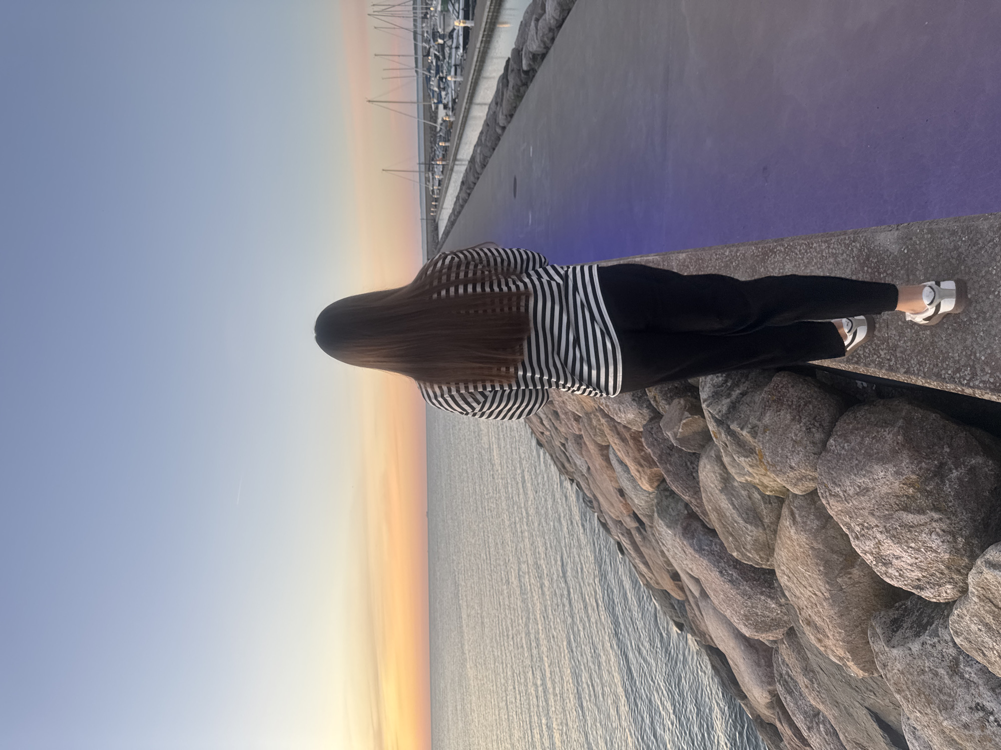A full shot, captured from behind, shows a person standing on a stone embankment with a paved walkway to their right. The person, facing away from the camera, has long, dark hair and is wearing a black and white horizontally striped long-sleeved shirt and black pants. White shoes with black accents are visible on their feet. To the left of the embankment is a body of water, with the horizon line marked by a soft orange and yellow sunset. To the right of the walkway, a marina with numerous sailboats is visible. The walkway itself is illuminated with a soft purple light. The sky transitions from a pale blue at the top to the warmer sunset colors near the horizon. The location is identified as Tallinn, Estonia, specifically the Pirita Marina.