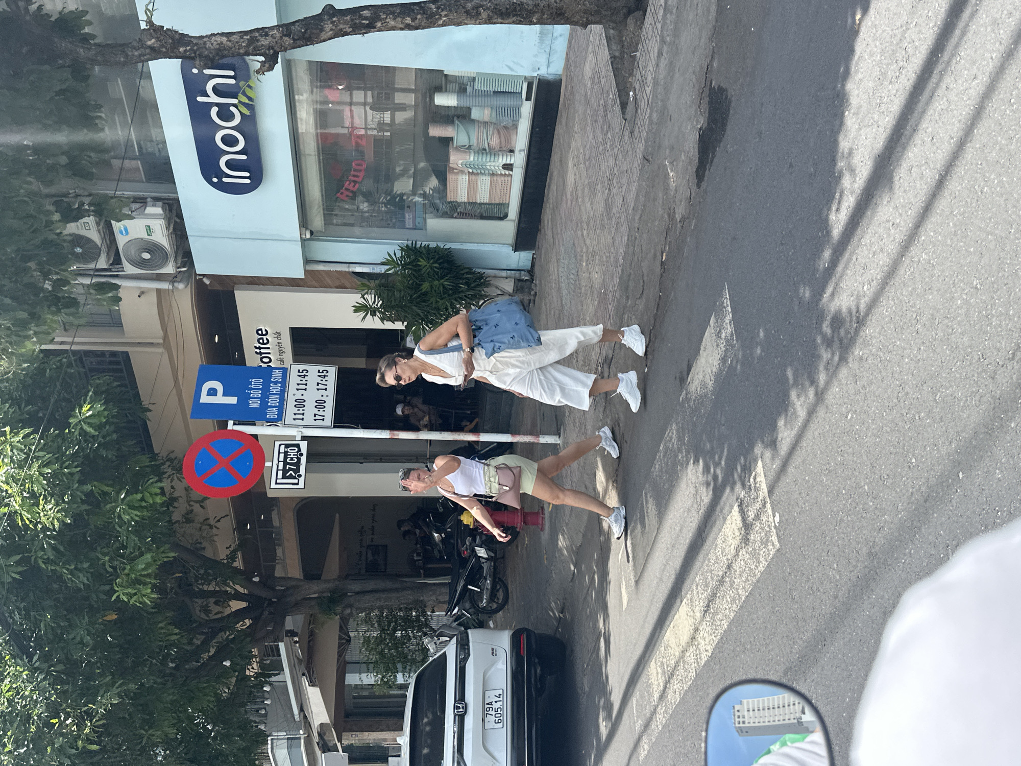 A street scene in Nha Trang Ward, Vietnam features two women crossing a street at a pedestrian crossing. The woman on the left, wearing a white tank top and light green shorts, is facing the camera and waving. She carries a light pink purse. The woman on the right, wearing a white jumpsuit, is walking away from the camera. She carries a large blue bag. In the background, several businesses are visible, including one with a sign that reads "inochi". The street is paved, with a white pedestrian crossing marked. A white SUV with license plate "79A 605.14" is parked on the left side of the street. Trees line the sidewalk, and there is a red fire hydrant near the pedestrian crossing. The sky is blue with some clouds.
