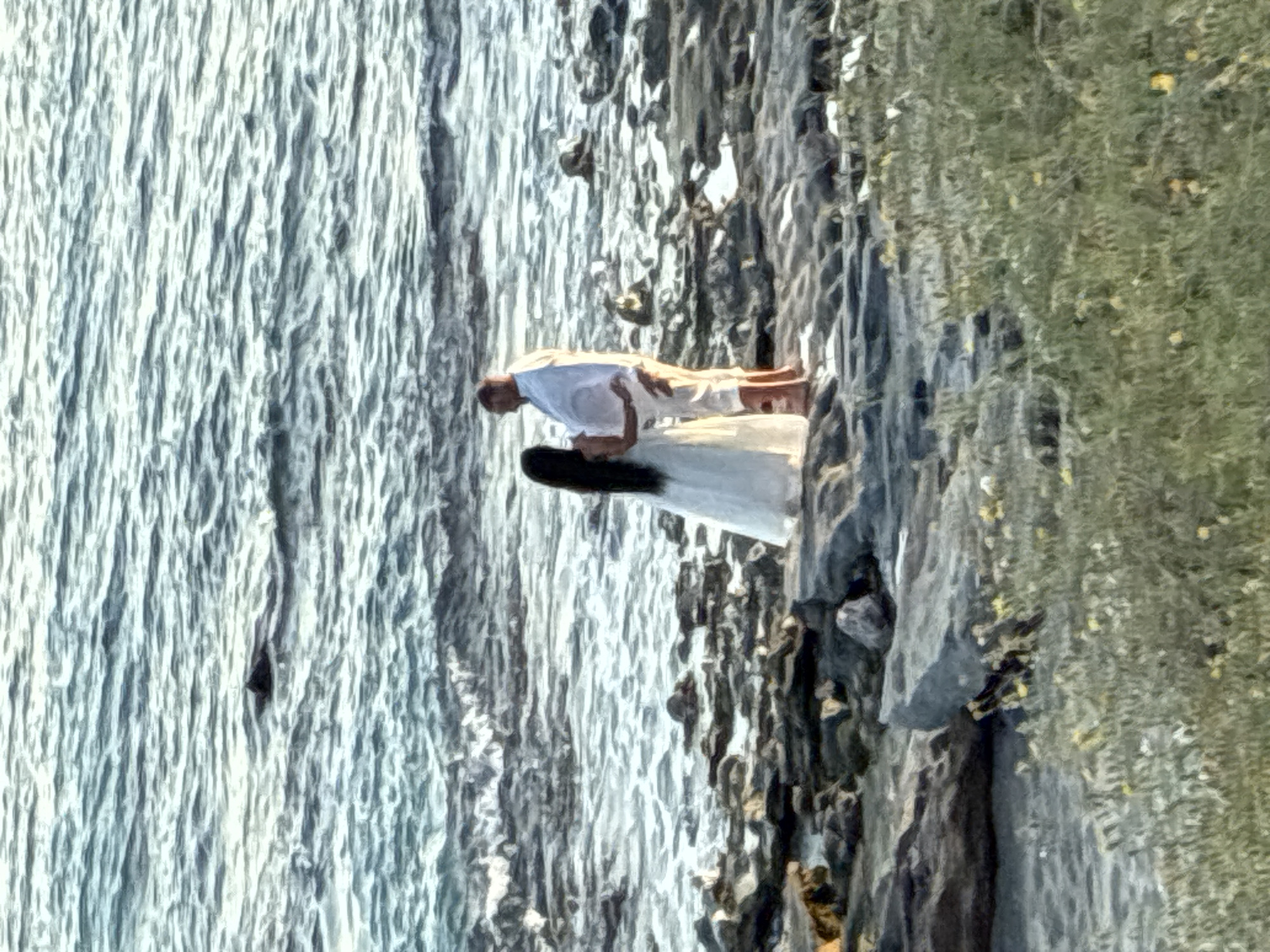 A couple stands on a rocky shore with the sea in the background. The woman on the left wears a long white dress and the man on the right wears a white shirt and light-colored shorts. They are facing each other, and the man has his arm around the woman. The sea is a dark blue with white waves. The shore is made of dark rocks. In the foreground, there is green grass and some small yellow flowers. The scene is lit by natural light, suggesting it is daytime. The location is not definitively Alanya, Turkey, as no specific landmarks are visible.