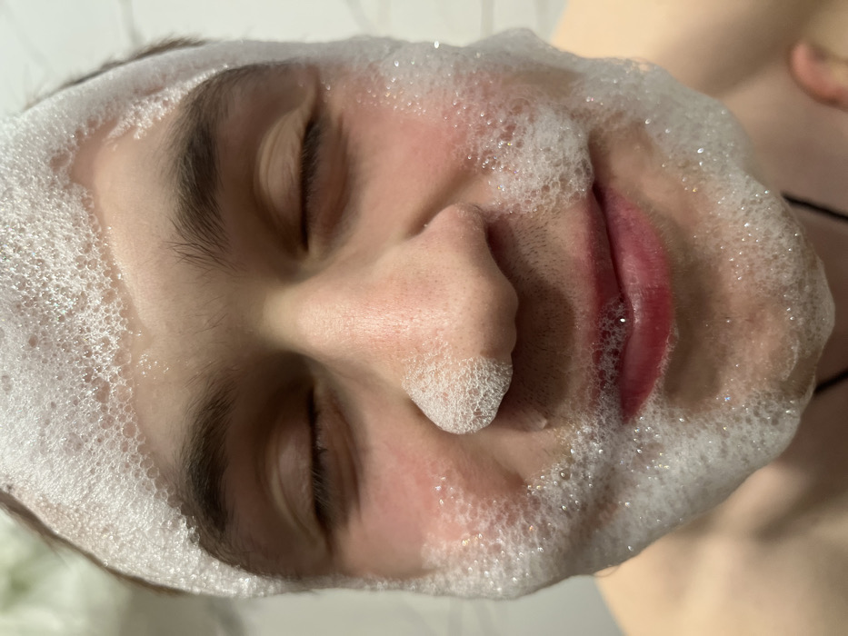 A close-up, rotated image shows a person's face covered in white foam or bubbles. Their eyes are closed, and the foam clings to their eyebrows, nose, cheeks, and lips. The person's skin is fair, and some redness is visible on the cheeks. Some bubbles appear to be touching or entering the open mouth. The background is blurry and includes a hint of white tiles and a shoulder.