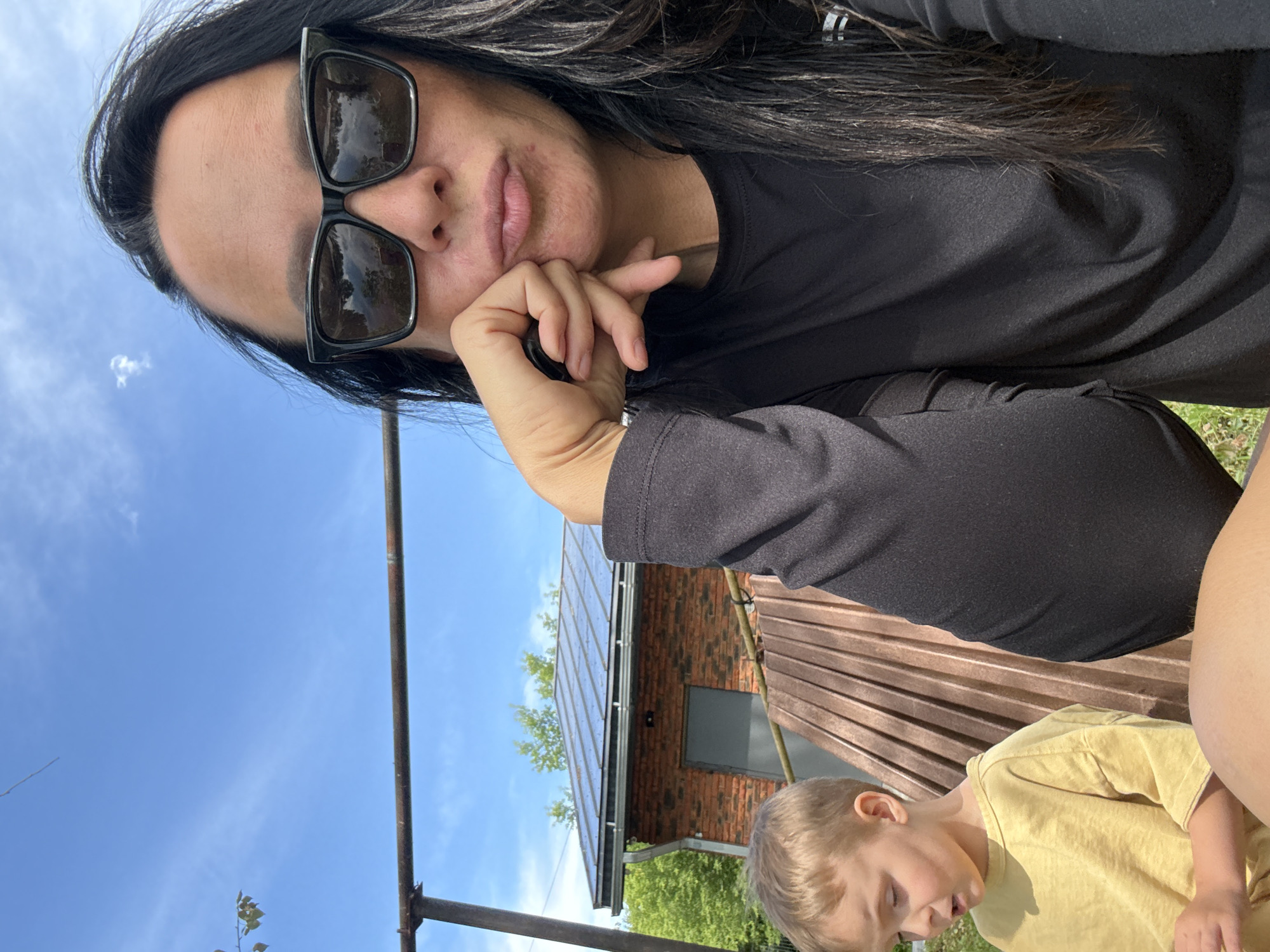 A young boy in a yellow shirt sits next to a woman wearing sunglasses and a black long-sleeved shirt. The woman rests her chin on her hand, and her lips are slightly parted. Behind them is a red brick building with a metal roof and trees. The sky is blue with scattered clouds. The scene appears to be outdoors, possibly in a backyard or rural area. The time of day is likely daytime, given the bright sunlight. The location is identified as Chaikovsky, Russia.