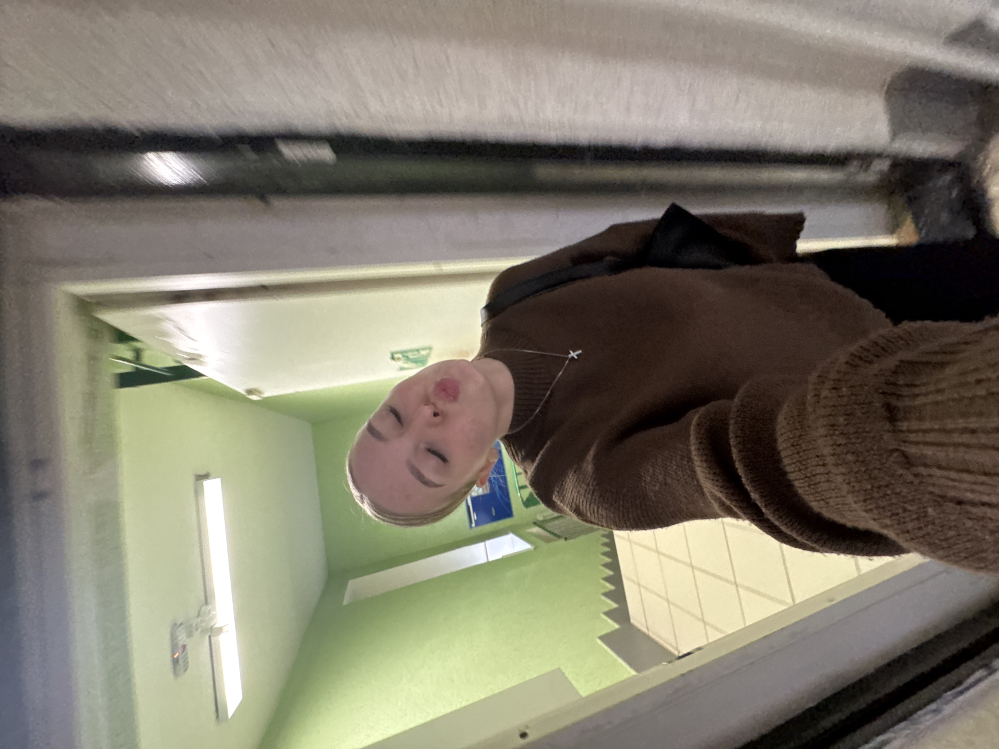 A woman is inside a building in Perm, Russia. The building has a green hallway with a long rectangular light fixture on the left side. The hallway floor is white tile. The woman is wearing a brown sweater and a black bag. She is looking at the camera and pouting.