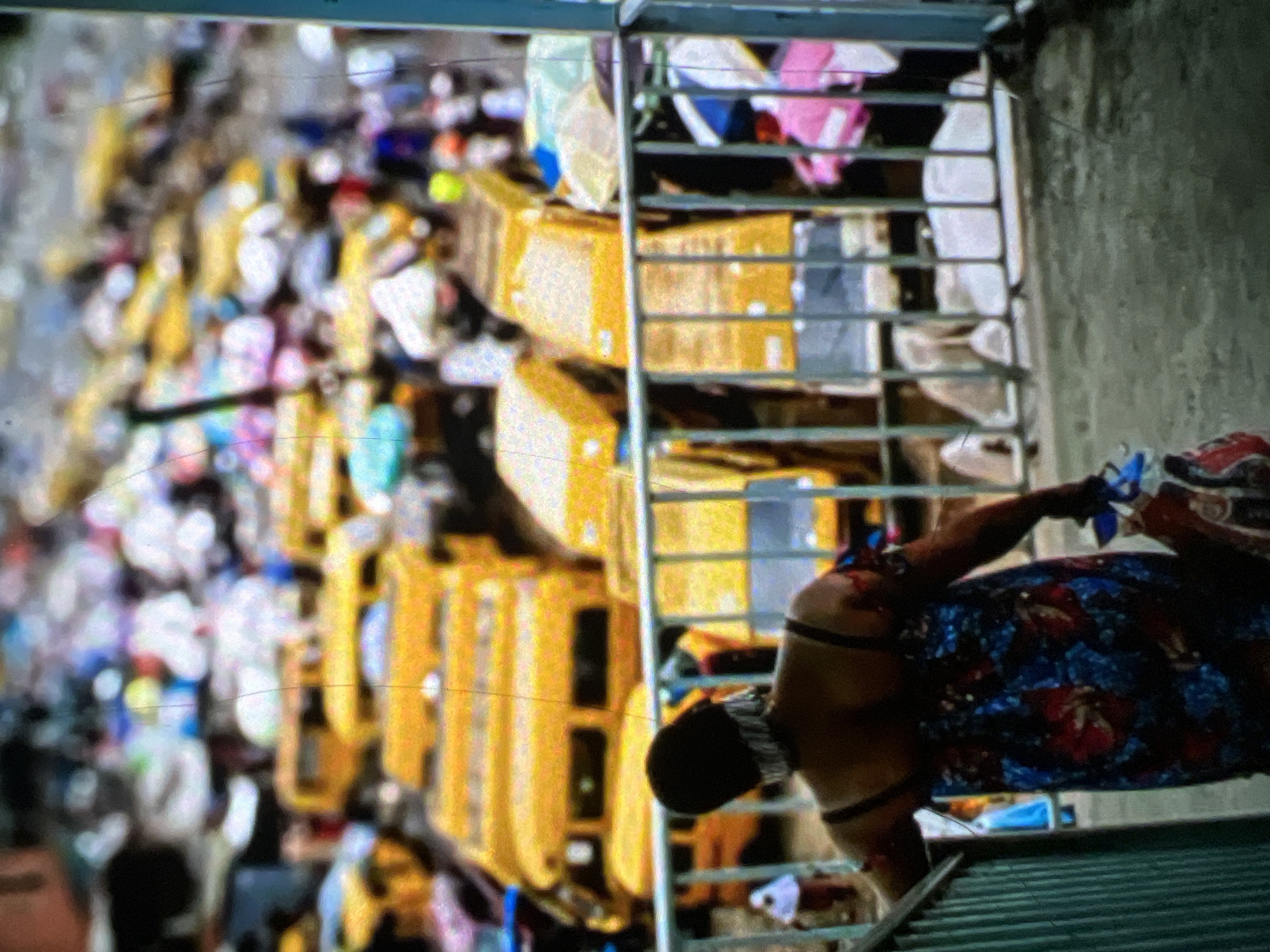The image shows a crowded scene in Lagos, Nigeria, likely a busy transportation hub or market. In the foreground, a woman with dark hair and wearing a colorful floral dress is walking down a staircase, holding a bundle of items. Her back is to the camera, and she appears to be wearing a headwrap or embellished hair accessory.
Behind the woman, a dense collection of yellow buses, possibly minibuses or commercial vehicles, are parked closely together. The buses are a dominant visual element, suggesting a significant volume of public transportation or a large fleet. The background is blurred, but it indicates a bustling urban environment with many people and vehicles. The overall impression is one of vibrant activity and the characteristic energy of Lagos. The scene appears to be outdoors, possibly near a road or a terminal. The presence of what looks like a metal railing on the left side of the image further suggests an elevated vantage point, perhaps a pedestrian overpass or walkway overlooking the street and the parked buses. The image is somewhat grainy, which may obscure finer details.