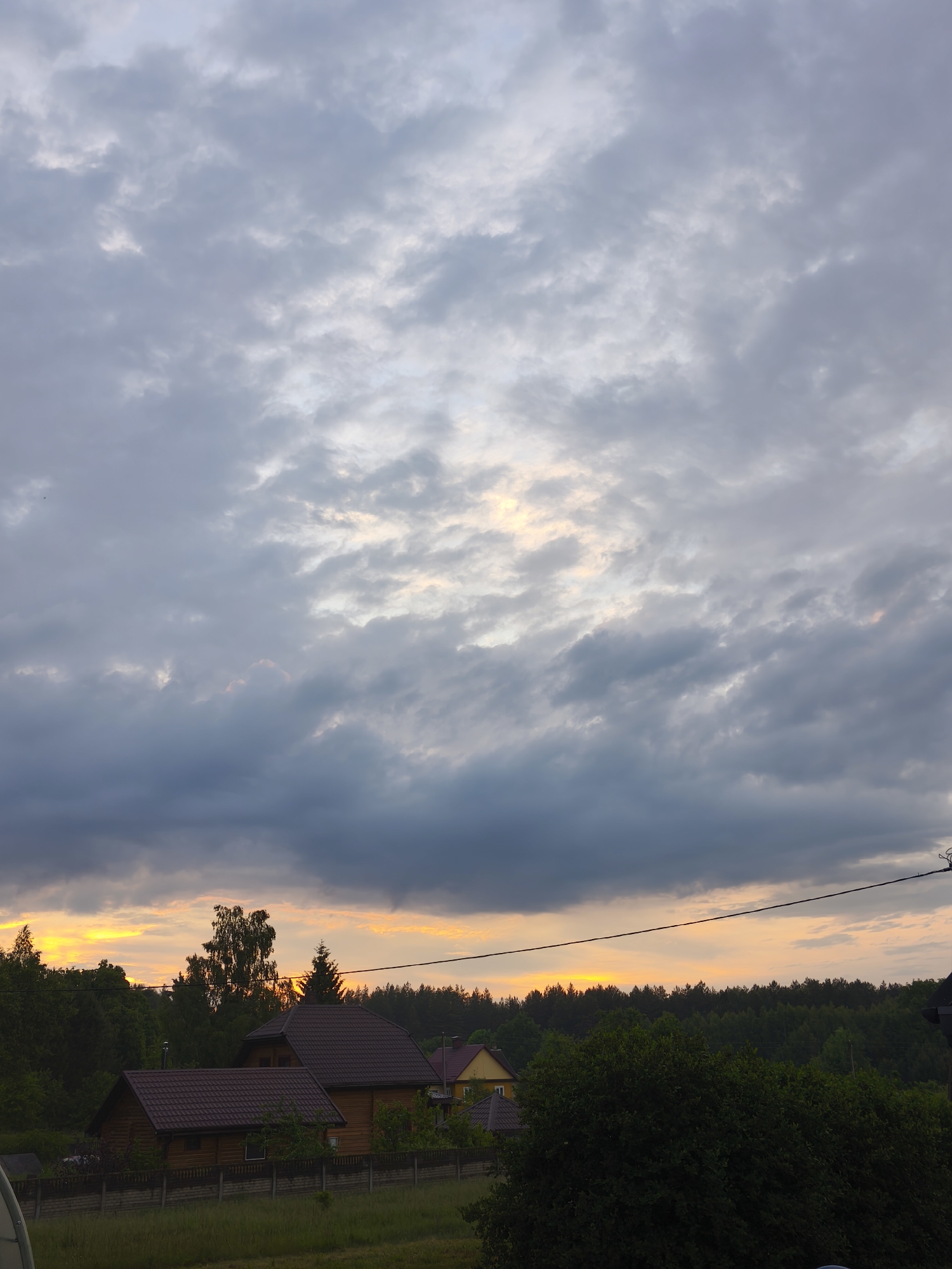 The image depicts a rural or semi-rural landscape in Парэчча, Belarus, at either sunrise or sunset. The upper two-thirds of the frame are dominated by an overcast sky composed of multiple layers of gray and off-white clouds, with patches of diffuse light indicating a hidden sun. Directly above the horizon, a distinct horizontal band of intense orange and yellow light is visible, suggesting the sun's low position.
In the lower third of the image, a dense line of dark green deciduous and coniferous trees forms a silhouette against the illuminated horizon, indicating a forested area. Below the tree line, multiple residential structures are present. In the mid-left, two prominent wooden houses with dark brown, multi-pitched roofs are visible; one larger structure with a smaller adjacent building, both appearing to be log cabins. A partial view of a yellow-sided house is present further right, partially obscured by trees.
A low, light-colored fence, possibly concrete or stone, runs horizontally in the mid-foreground, separating a grassy area from the houses. Lush green vegetation, including bushes and trees, populates the midground. A dark electrical wire or cable extends horizontally across the upper right portion of the midground. In the bottom-left corner, a white, parabolic dish-like object is partially visible. No individuals or overt human activity are depicted.