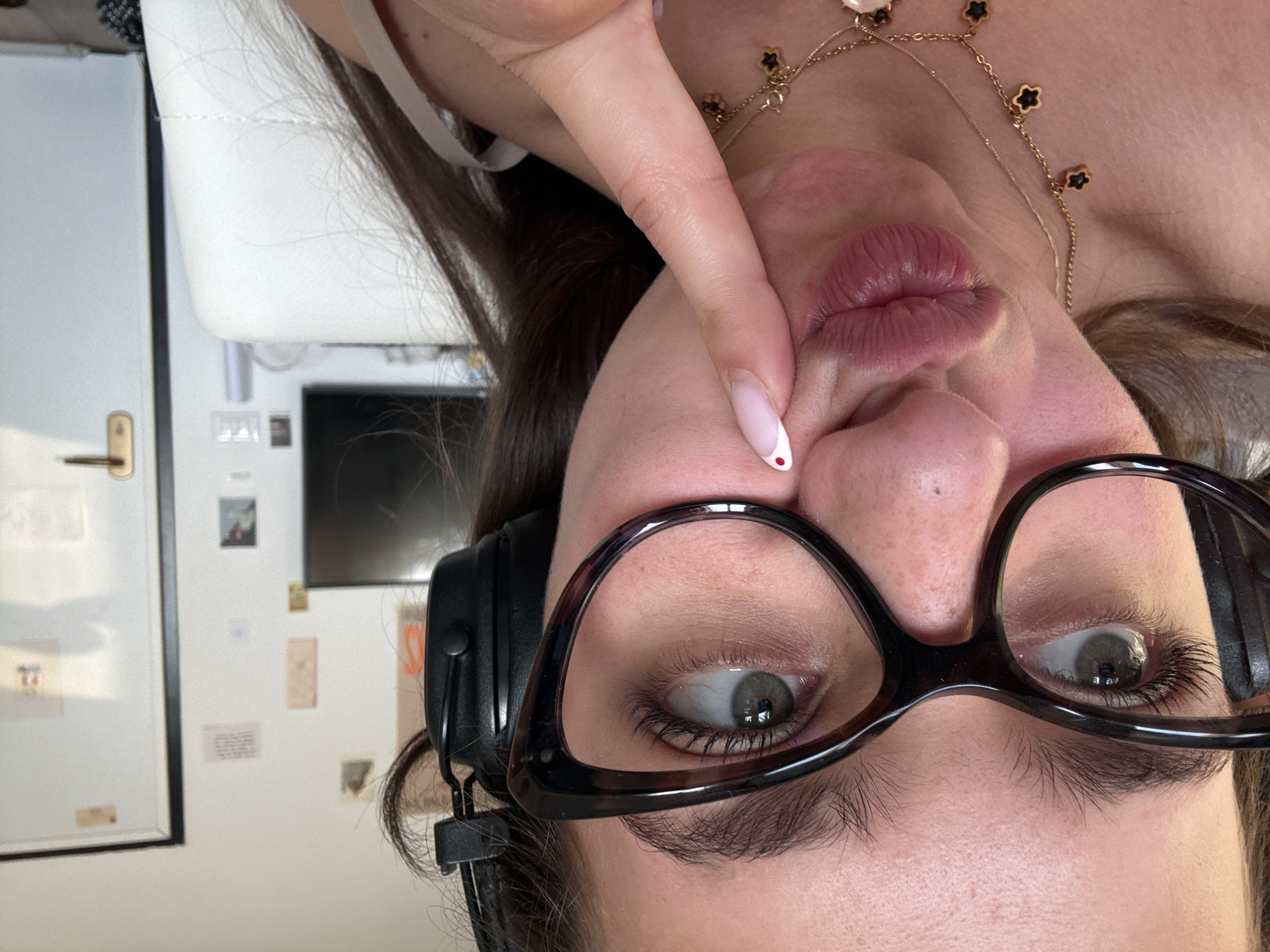 A close-up, inverted, and slightly skewed perspective of a person's face. The subject has brown hair, wears black-rimmed glasses, and black headphones are partially visible on the side of their head. The person's lips are pursed, and they are touching their cheek with a finger that has a light pink nail polish with a red dot on the tip. A gold necklace with clover-shaped pendants is visible. In the background, there is a white door with a gold handle, a framed picture, a computer monitor, and several small papers or images on the wall. The setting appears to be indoors. There is no clear indication that the location is Milan, Italy.