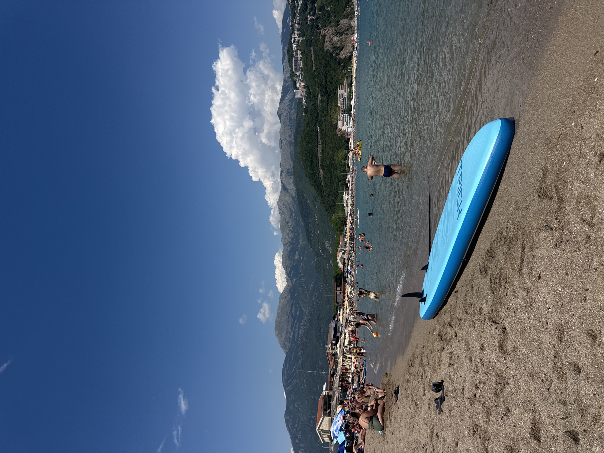 The image depicts a crowded beach scene on a sunny day, likely in Prijevor, Montenegro, with mountains in the background. In the foreground, a bright blue paddleboard rests on the sandy shore near the water's edge. The water is clear and shallow, with several people wading and swimming. Further back, the beach is lined with umbrellas and sunbathers. The backdrop features lush green hillsides dotted with buildings, and prominent mountains rise behind them under a clear blue sky with scattered white clouds.