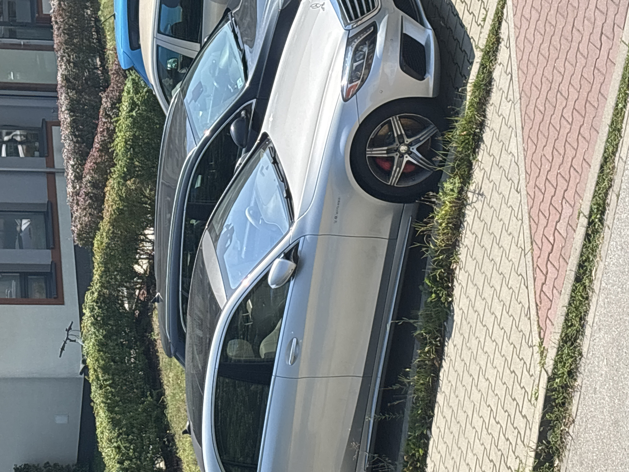A silver Mercedes-Benz sedan with a black panoramic sunroof is parked on a paved surface in front of a building. The car's wheels have five spokes and red brake calipers. A black SUV is parked behind it, and a blue vehicle is visible further in the background. The scene is outdoors, with a hedge and grass in the foreground. The paving stones are a light gray interlocking pattern with a section of reddish-pink pavers. The image appears to be taken on a sunny day. The location is in Krakow, Poland, based on the context provided. There are no people visible in the image.