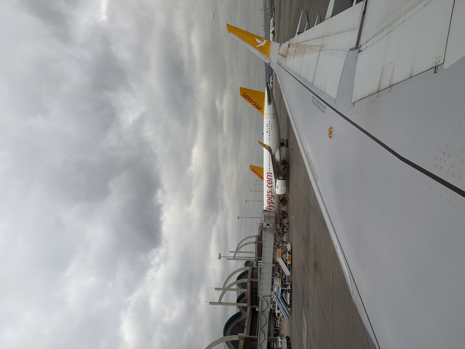 This image depicts an airport apron scene under an overcast sky, viewed from an aircraft window overlooking its wing. The location is Sabiha Gökçen International Airport (SAW), situated near Tuzla, Istanbul, Turkey.
In the foreground, the light gray, metallic wing of a commercial airliner, from which the photograph is taken, extends across the lower right frame. The wing displays visible structural lines, panel seams, and numerous small, circular water droplets on its surface. A yellow rectangular safety marking is affixed to the wing.
In the midground, two white-bodied Pegasus Airlines aircraft are parked on the asphalt tarmac. The closer aircraft, with a yellow vertical stabilizer featuring a white Pegasus logo, displays "flypgs.com" in red and "PEGASUS" in yellow on its fuselage. This aircraft is connected to a passenger jet bridge that extends from the terminal building. Surrounding the aircraft are various ground support vehicles, including a blue and white mobile passenger stair unit, yellow baggage carts, and a small yellow tug vehicle. Several individuals, consistent with airport ground crew, are visible near the aircraft and jet bridge, engaged in operational activities. A second, identical Pegasus Airlines aircraft is parked further behind the first, partially obscured.
The background features the modern terminal building of Sabiha Gökçen International Airport, characterized by its distinctive arched support structures and extensive glass panels. Partially visible text on the terminal structure reads "INTERNATIONAL AIRPORT." Further in the distance, other aircraft tails and airport infrastructure are faintly visible. The sky above is entirely covered by dense, grey stratocumulus clouds. A tiny, distant aircraft is observable in the upper right quadrant of the sky.