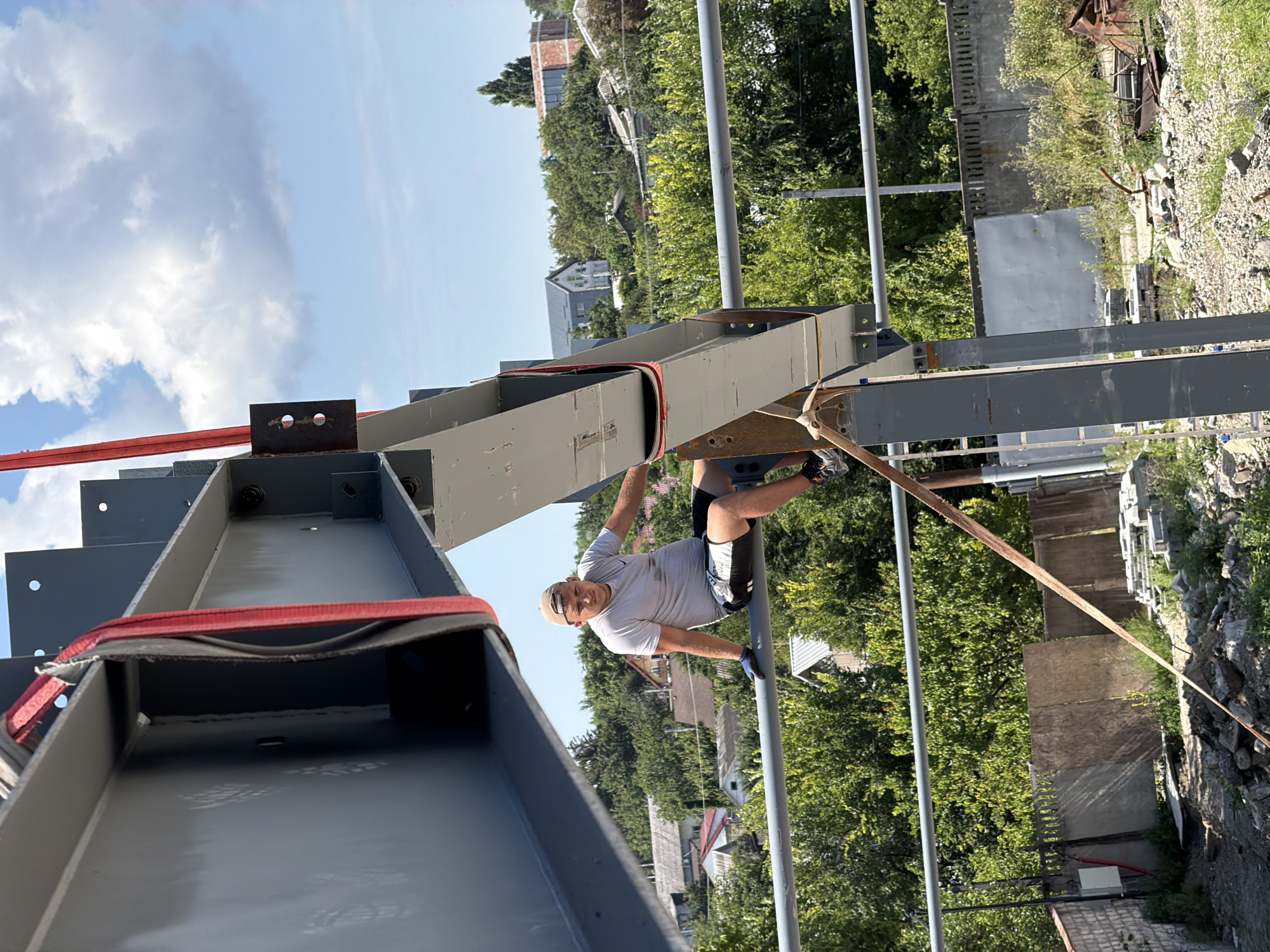 A man in a grey t-shirt, black shorts, a beige baseball cap, and blue work gloves is sitting on a horizontal metal beam. The beam is part of a large steel construction, painted grey, and appears to be suspended by red and black straps. The man is looking towards the camera. In the background, there are trees, houses, and a blue sky with white clouds. The setting appears to be an outdoor construction site. Based on the architecture and vegetation, the location is likely Vatra, Moldova.