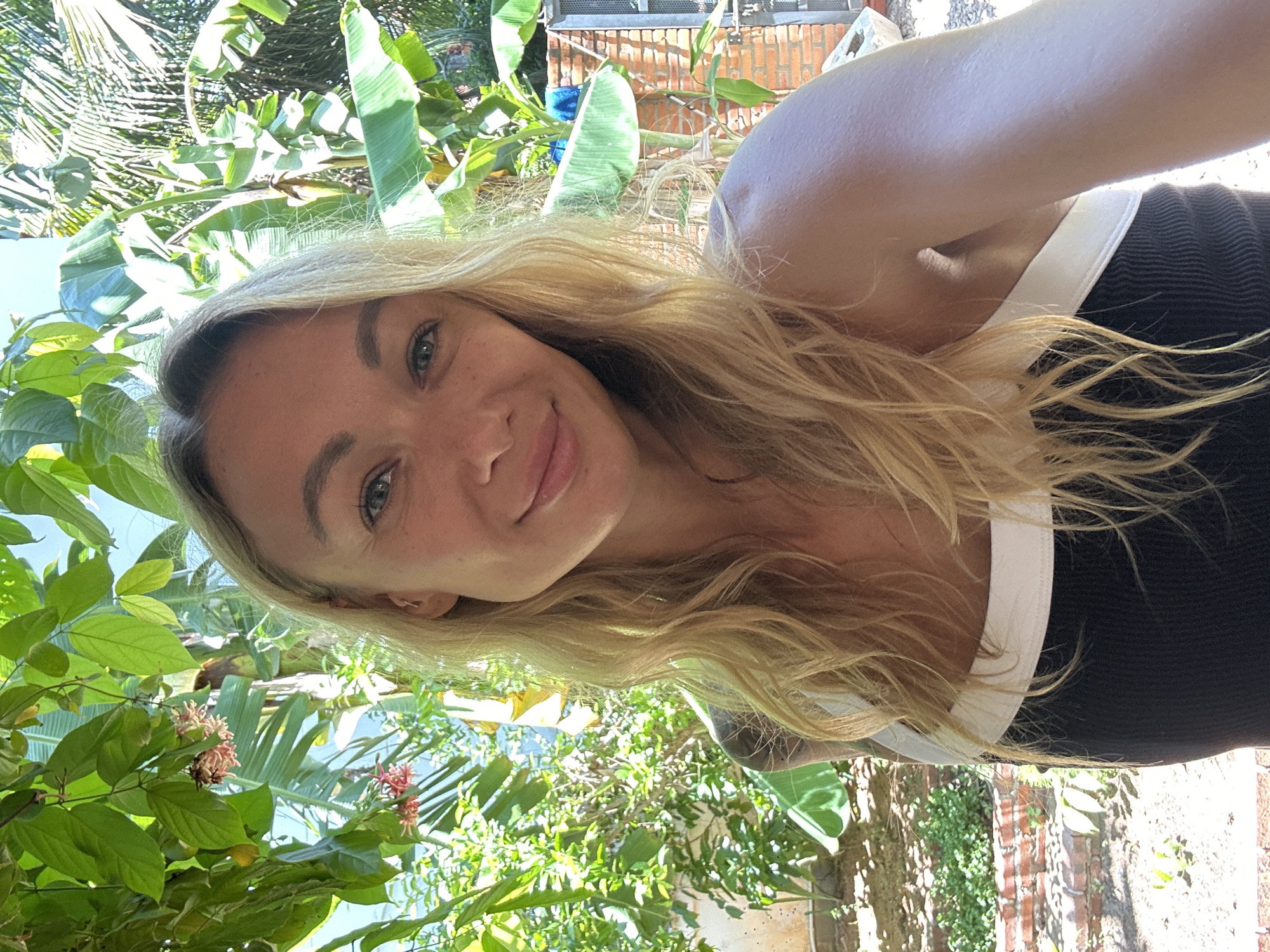 A blonde woman with blue eyes smiles at the camera. She is wearing a black and white tank top. She has a tattoo on her left arm. She is outdoors, with green foliage and a brick wall in the background. The scene appears to be in a tropical location, possibly Mũi Né, Vietnam, given the lush greenery and sunlight.