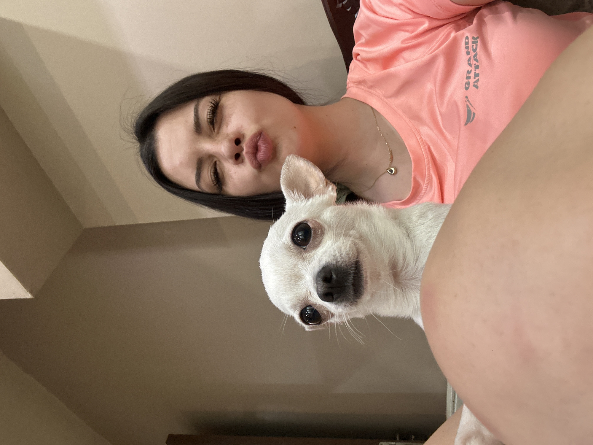 A young woman with dark hair and freckles is taking a selfie with a white chihuahua. She is making a kissing face with her eyes closed. She is wearing a coral-colored t-shirt with a logo that reads "GRAND ATTACK" and a delicate necklace. The chihuahua is looking directly at the camera with large, dark eyes. The background appears to be an indoor setting with neutral-colored walls. The image was taken in Sliven, Bulgaria.