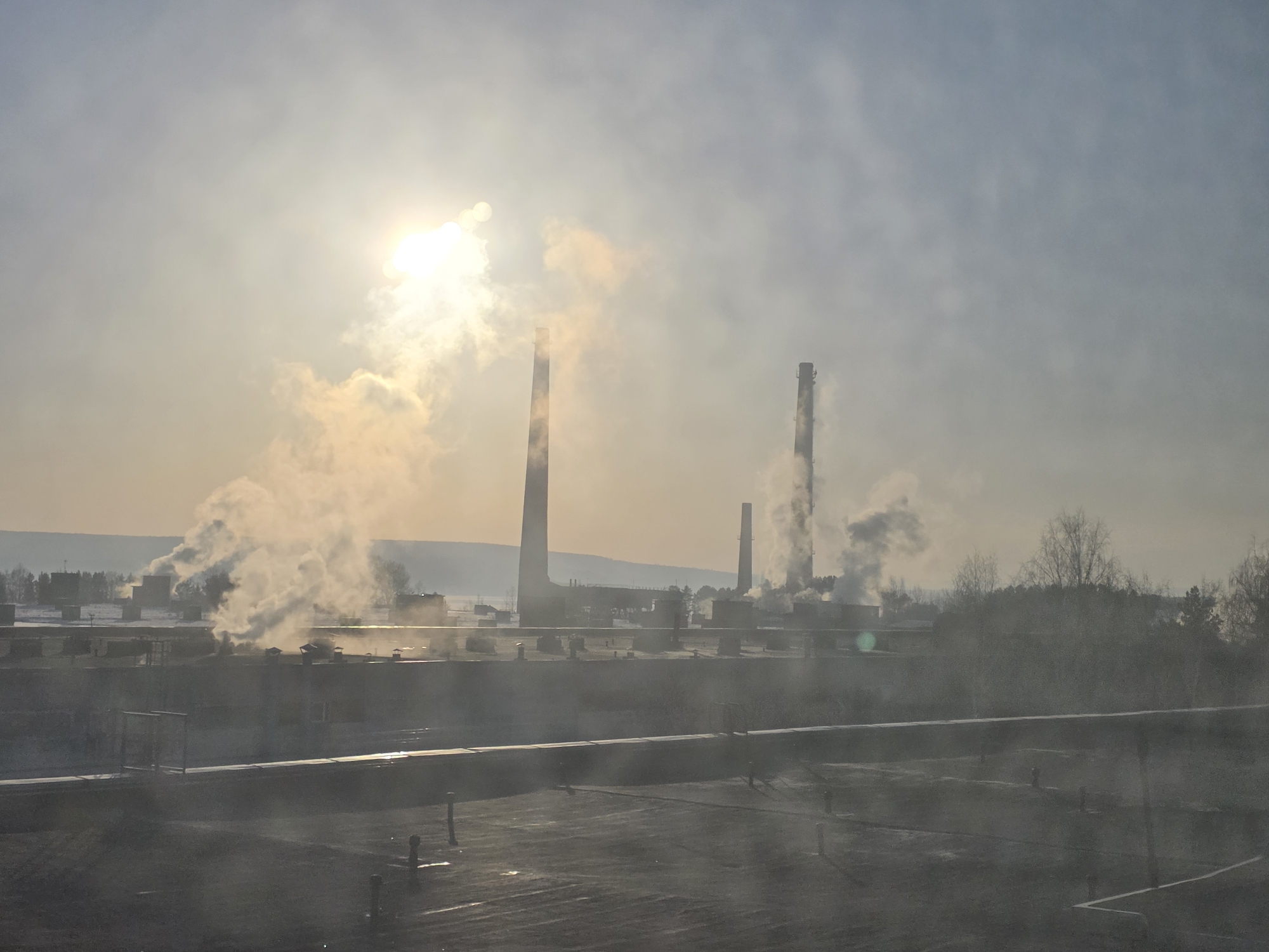 The image depicts an industrial scene, potentially in Chaikovsky, Russia, given the likelihood from the surrounding dataset context. There are multiple factory chimneys or smokestacks emitting plumes of smoke or steam, suggesting industrial activity. The smoke partially obscures the sun, creating a hazy atmosphere. A network of industrial buildings is visible, partially obscured by the emissions. The foreground appears to show a flat surface, perhaps a rooftop or pavement, with some linear features and minimal detail. The overall impression is one of industrial presence and activity with potential environmental implications. The horizon shows trees and low rolling hills or distant buildings. There are no visible people.
