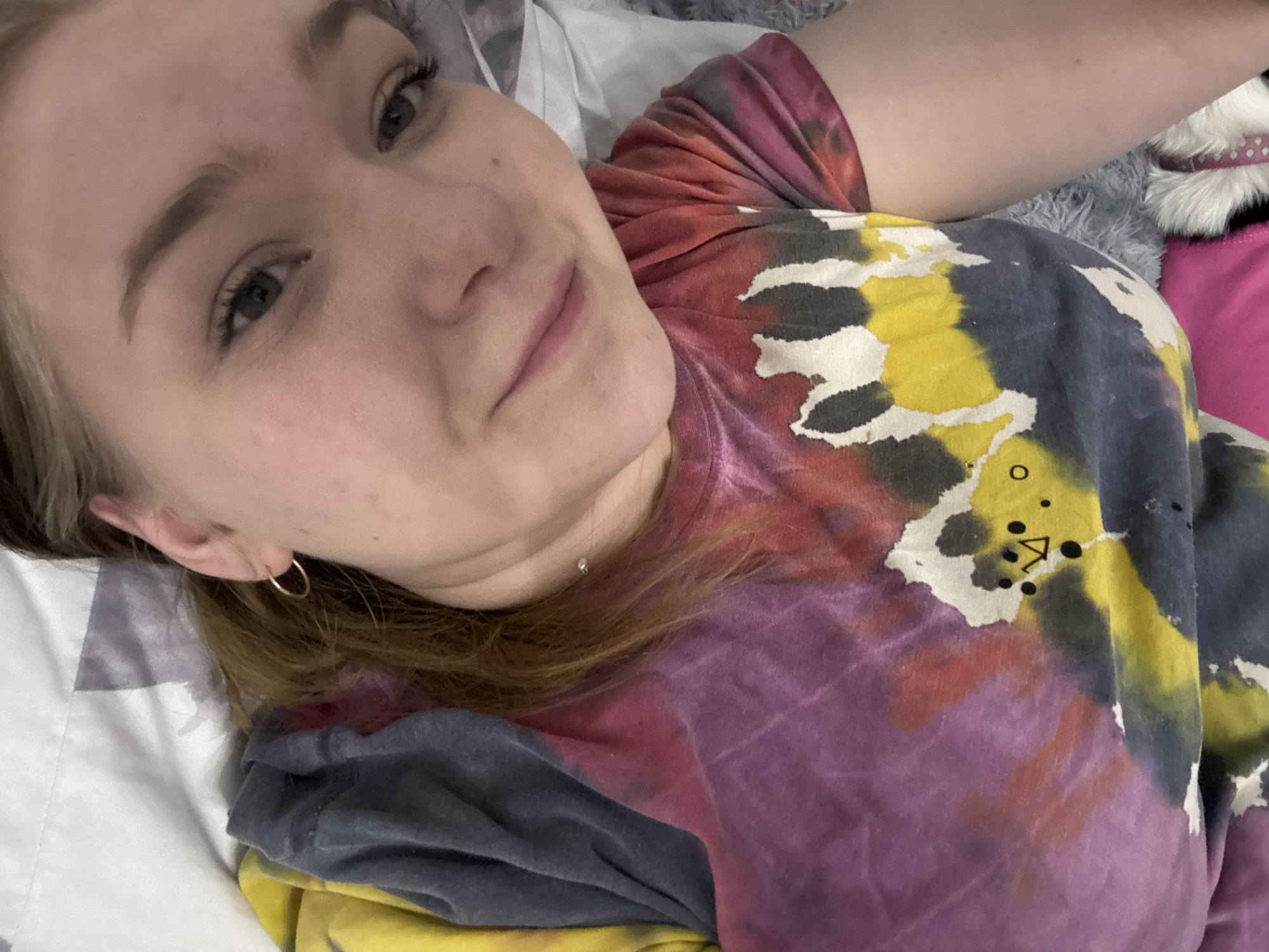 A woman lies on a bed with patterned bedding. She is wearing a tie-dye shirt with shades of red, pink, yellow, and gray. Her hair is light brown, and she has a gold earring in her left ear and a small necklace. A furry object with a pink collar is visible in the upper right. The location is not apparent from the visual information in the image and cannot be confirmed as Chaikovsky, Russia.