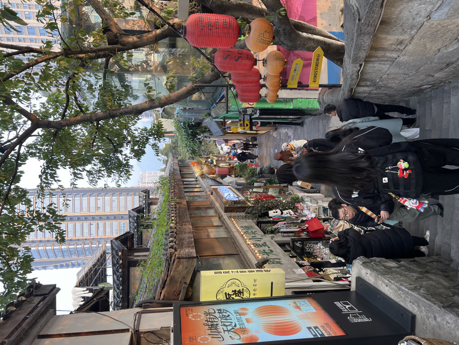 The image depicts a steep, stepped pedestrian alleyway located within the Yuzhong District of Chongqing, China. The scene is illuminated by natural daylight, possibly late afternoon or early morning, indicated by soft shadows and warm highlights.
In the foreground, a female subject with long dark hair, wearing a black long-sleeved top and black trousers, is descending a gray stone staircase. She carries a black backpack adorned with several small, colorful decorative keychains. To her left, slightly behind, a male subject in a dark jacket with a multi-colored strap over his shoulder holds a mobile phone. Adjacent to them on the left, another female subject in a dark winter coat faces a shop display.
The alleyway is bordered on both sides by multi-story buildings constructed in a traditional Chinese architectural style, featuring dark tiled roofs, some with visible green foliage growing upon them, and wooden structural elements. Shopfronts line both sides of the alley, displaying merchandise and illuminated signs with Chinese characters. On the left, a large, vertical electronic display board shows an illustration of orange lanterns and Chinese text. Higher up on the left, a prominent gilded rooster or phoenix statue is mounted above a shop entrance.
On the right side of the alley, a substantial tree with green leaves stands beside the staircase. Numerous red and orange paper lanterns, some inscribed with Chinese characters, are suspended from its branches and across the alley. A yellow directional sign attached to the tree points towards "BANDSTAND" and a Chinese character location. The right side of the staircase is bordered by a low gray stone wall.
In the mid-ground and background, approximately 15 to 20 individuals are visible, primarily walking down the steps or observing the shop displays. The alley continues to slope downwards into the distance. Beyond the traditional buildings, modern high-rise structures with glass facades are visible against a light sky, establishing a contrast between historical and contemporary urban development. Distant hills or mountains are faintly discernible on the horizon.