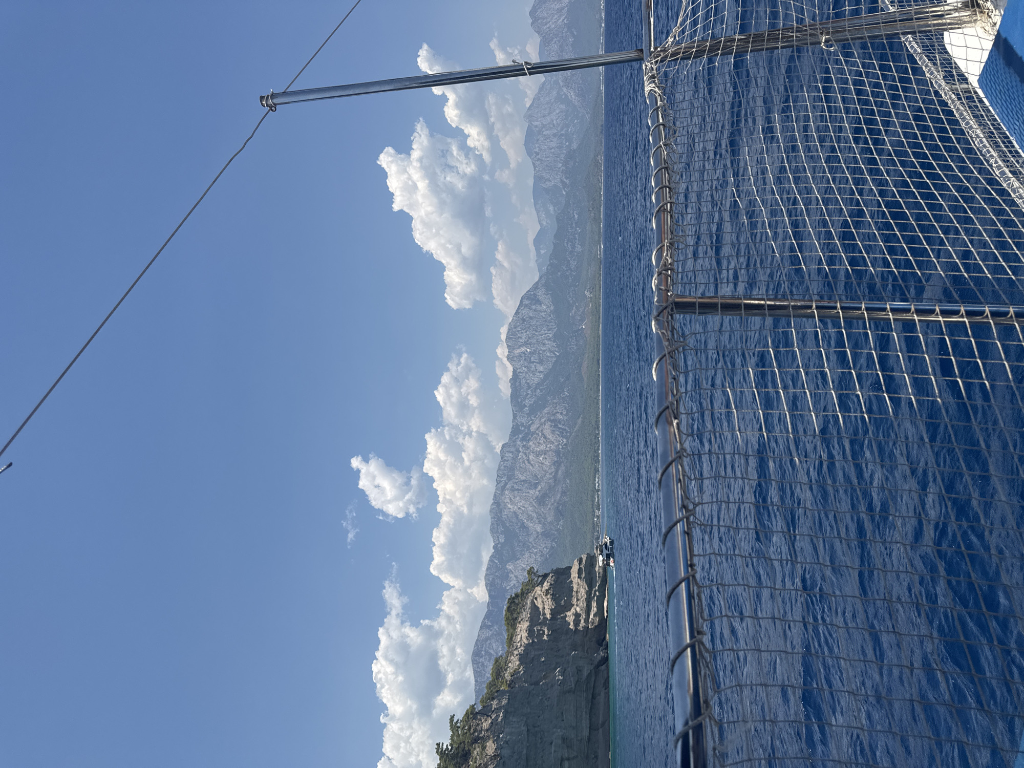This is a photograph taken from a boat on a sunny day, looking towards the coast. The sea is a deep blue, with gentle waves. In the foreground, a metal railing with netting is visible, suggesting the perspective is from the deck of a vessel.
The coastline features a dramatic, rocky cliff face on the left, covered in green vegetation at its peak. Beyond the cliff, a small white boat with people on board is visible. The water near the shore is a lighter turquoise color.
In the distance, a range of majestic, rugged mountains forms a backdrop to the scene. Their slopes are covered in a mixture of rock and sparse vegetation, and their peaks are sharp and angular. The sky above is a clear, bright blue, dotted with fluffy white cumulus clouds. The overall impression is one of a serene coastal landscape in a Mediterranean setting. This type of scenery is characteristic of the coastal areas around Kemer, Turkey, known for its dramatic cliffs, clear blue waters, and mountain backdrops.