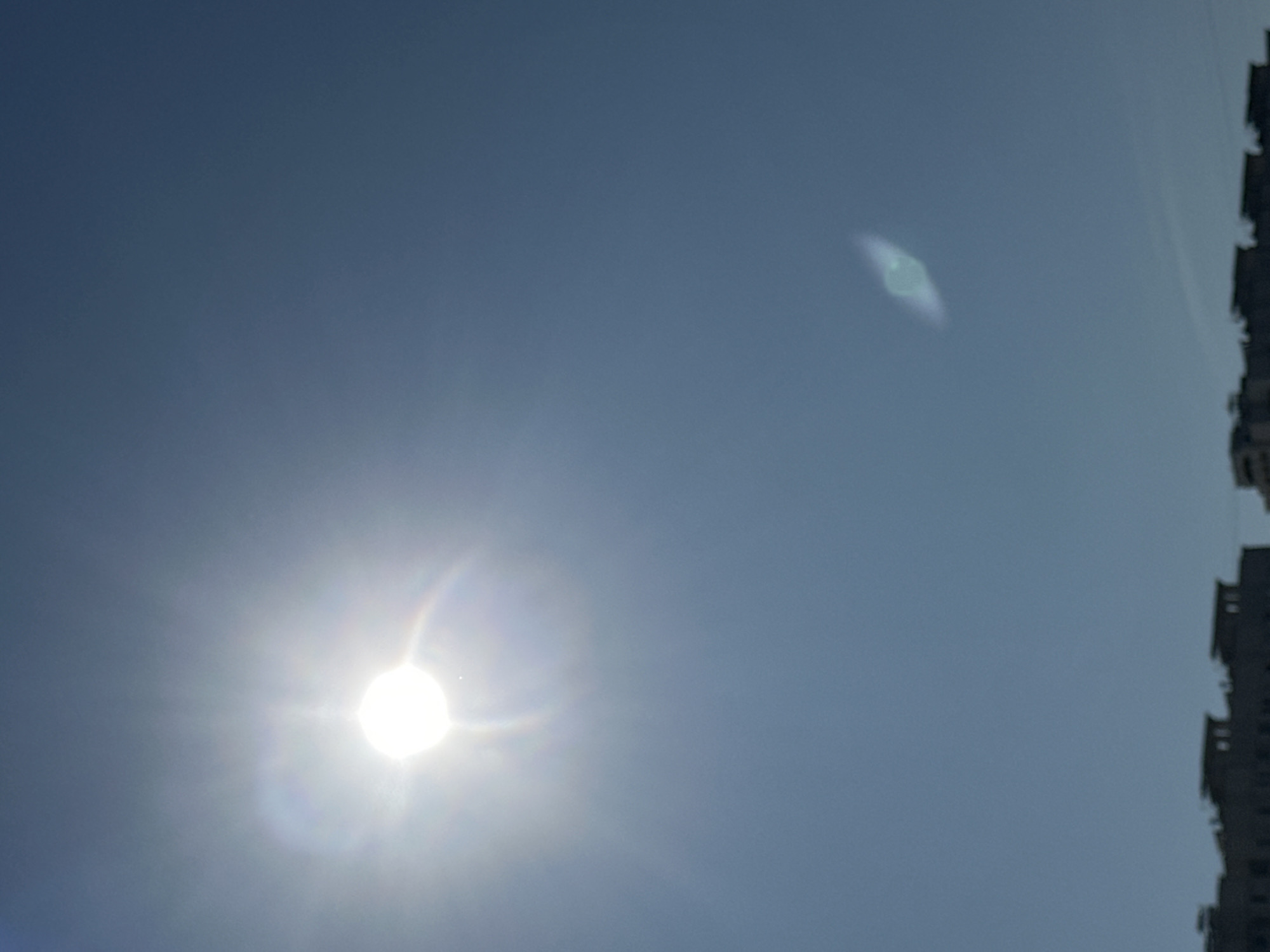 The image depicts a clear blue sky above Podolsk, Russia. The sun is prominently visible in the upper left quadrant, emitting bright rays. A lens flare, appearing as a faint, elongated greenish shape, is present in the middle right of the sky. Silhouetted against the sky in the lower portion of the image are the tops of several buildings, suggesting an urban environment. The architecture appears to be residential or commercial, with flat roofs visible. The overall impression is of a bright, sunny day in the city.