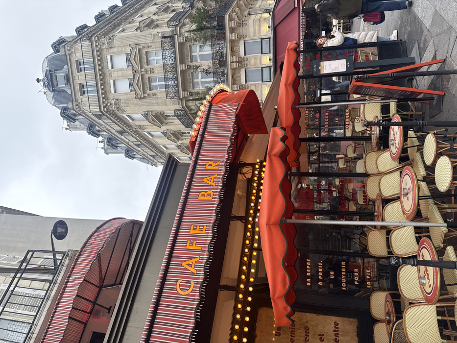 The image depicts an outdoor Parisian café in the Les Halles district, near Église Saint-Eustache, Paris, France, under an overcast sky. The café occupies the ground floor of a building, featuring prominent red and white striped awnings. The main awning displays "CAFE BAR" in yellow capital letters, with a string of illuminated round yellow lights beneath it.
An extensive outdoor seating area is furnished with numerous small, round bistro tables and light-colored wicker-backed chairs, many of which are stacked. Several large, circular red umbrellas are open over these tables. Small white ashtrays are present on some tabletops. Through the large glass windows of the café, additional indoor seating is visible, along with reflections of the surroundings. Blackboards or signs visible through the window display French text, including phrases like "UN REPAS À PARIS. UN SOUVENIR POUR LA VIE."
To the right of the café's entrance, a black sandwich board stands on the sidewalk, likely displaying a menu. Two individuals are present on the sidewalk near the café. One, further right, is seen from behind, wearing a dark jacket and blue jeans, carrying a red bag. The other, closer to the café, wears a long white puffer coat and white sneakers, looking away from the camera.
In the background, a grand, multi-story Haussmannian-style building dominates, constructed of light-colored stone. It features numerous windows, ornate wrought-iron balconies on several levels, and a distinctive mansard roof with dormer windows at its corner. A dark street lamp is attached to the upper part of the building on the far left. Dark bollards are visible in the foreground, bordering the seating area.