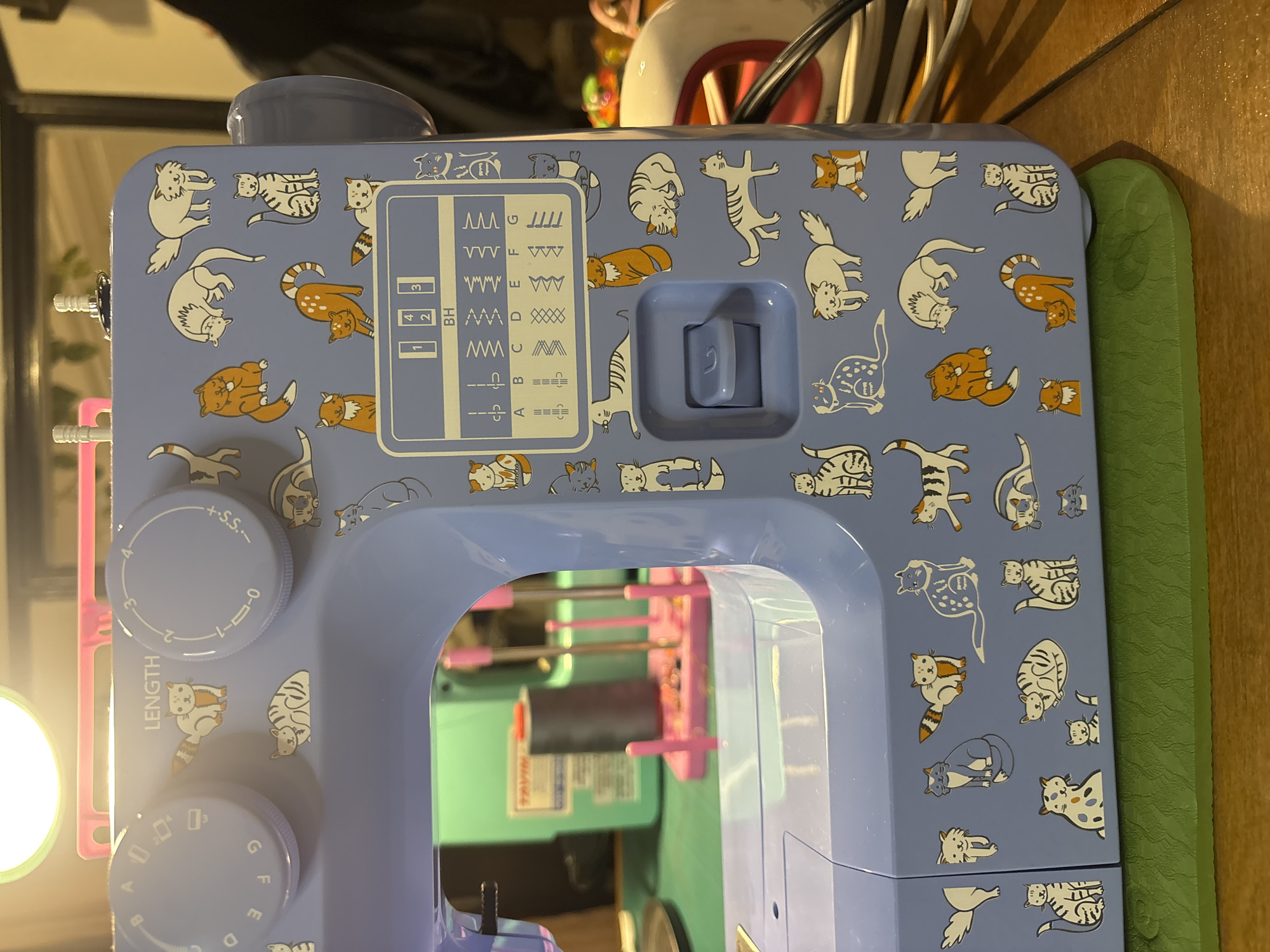 The image shows the top portion of a light blue sewing machine decorated with cartoon cats in various poses and colors, primarily white, orange, and gray. The machine features a stitch selection panel with labeled stitch types (A-G and 1-4 BH), a stitch length dial labeled "LENGTH" from 0 to 4 and "+S.S-", and a stitch width dial with letters A to G. The machine's internal structure is partially visible through the arm, revealing pink thread spools. The machine is set on a green surface next to a wooden one. The overall environment is a cluttered workspace, evidenced by other objects in the background.