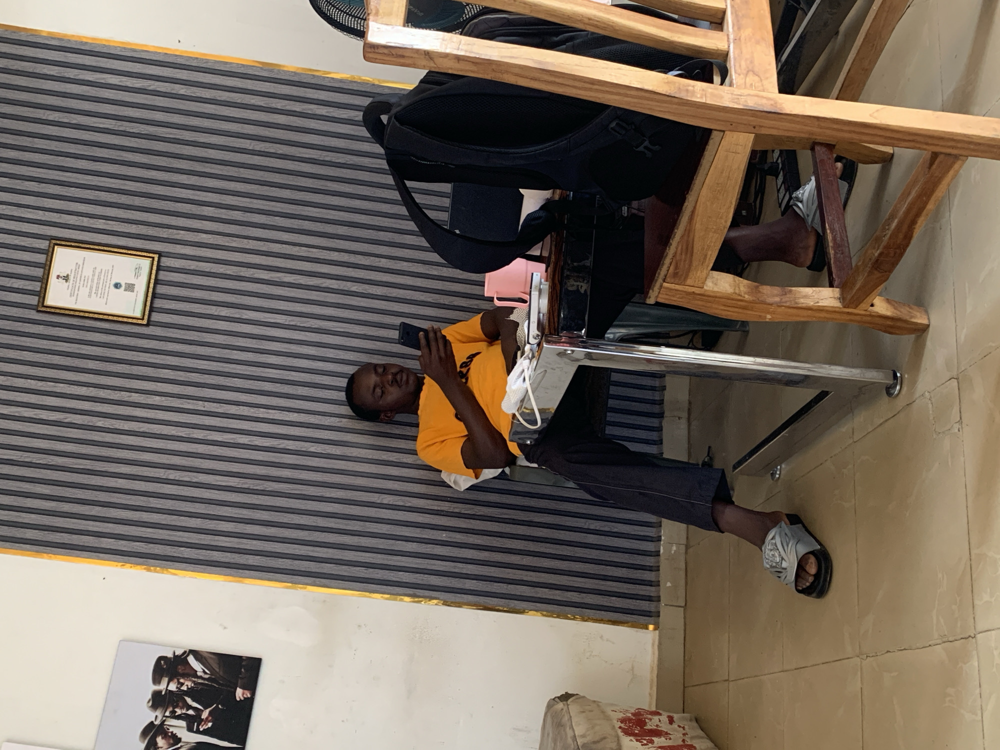 An indoor scene in Jos, Nigeria, shows a young man relaxing while using his smartphone. The man, wearing an orange t-shirt, dark trousers, and grey sandals, is seated in a wooden chair at a table. He is leaning back against a distinctive wall featuring horizontal grey and dark grey striped panels, with a gold trim running along the top where the wall meets the ceiling.
His left arm rests on the light-colored table, and a white face mask is draped near him, possibly on his lap or the table's edge. A black backpack is placed on the chair next to him. The table has a chrome or metallic frame. The floor is covered with light brown tiles.
On the striped wall, a framed certificate with a golden border is visible. While the text is unreadable, a crest or emblem is discernible at the top, and a QR code is visible near the bottom right of the document. Below the gold trim on the left, there's another image on a white wall, featuring people wearing hats, though details are indistinct. Part of a dark-colored fan can be seen in the upper right corner of the frame. The man appears calm and focused on his device.