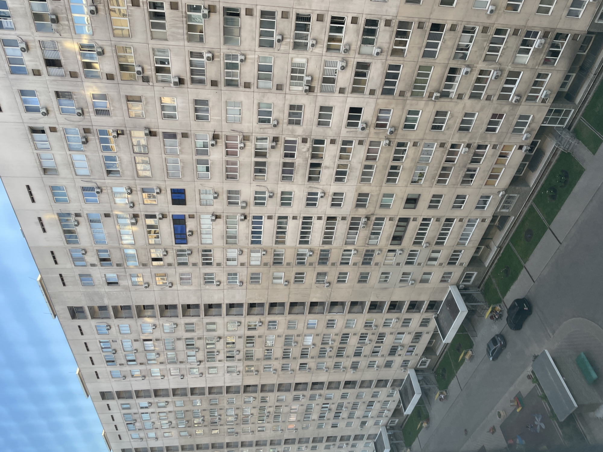 This media file captures an aerial view of a large, concrete apartment building in Almaty, Kazakhstan. The building features a grid of numerous windows and air conditioning units, suggesting a residential setting. The perspective is looking down and slightly to the side, revealing the side of the building and a portion of the surrounding area.
Below the building, a paved courtyard is visible with some green spaces and what appear to be children's play areas. There are a few cars parked in the courtyard, and some people are seated outdoors, possibly on benches or at tables. The scene appears to be during daylight hours, with bright sky visible to the left, indicating it is likely daytime. The weather seems fair, without any obvious signs of rain or overcast skies.
Notable details include the repetitive architectural style of the building, with many identical windows and AC units. The presence of parked cars and people in the courtyard suggests a normal day in a residential neighborhood. The overall impression is one of urban living in a city environment. No visible text is present in the image.