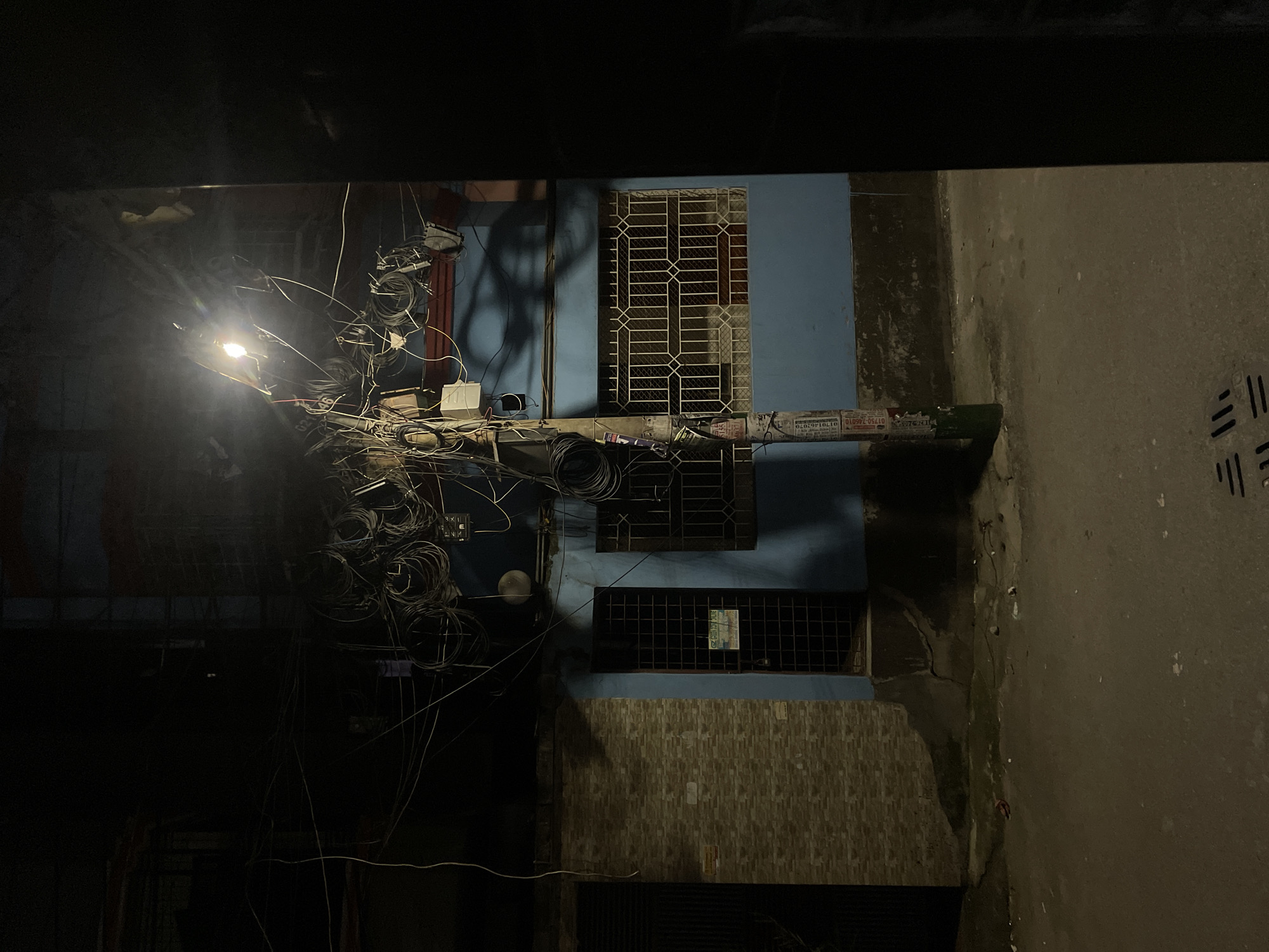 The image shows an urban alleyway at night, likely in Dhaka, Bangladesh, based on visual cues like building styles, power lines, and street conditions. Overhead, there is a chaotic web of electrical wires and equipment attached to a utility pole with a streetlight. These wires obscure a pale blue building facade with three barred windows of varying sizes. The building’s lower portion has tan-colored brickwork. The ground consists of worn concrete, stained with grime. A drainage grate is visible on the right side of the alley. The alley is dark, with most light emanating from the streetlight.