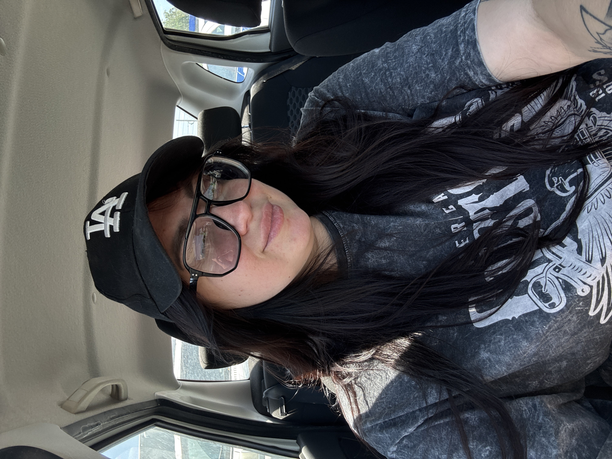 A female wearing a black LA baseball cap and black-framed eyeglasses, with long, black hair and a patterned grey t-shirt, is seated inside a vehicle. The car's interior includes grey upholstery. Outside, the daylight suggests daytime. The location is Unknown, Unknown.